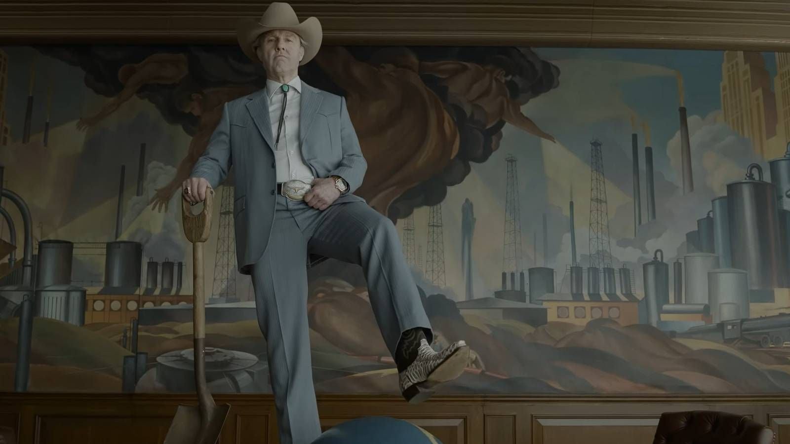 Ein Mann im Cowboyhut steht in Anzug und Cowboystiefeln vor einem Wandgemälde mit Industrielandschaft.