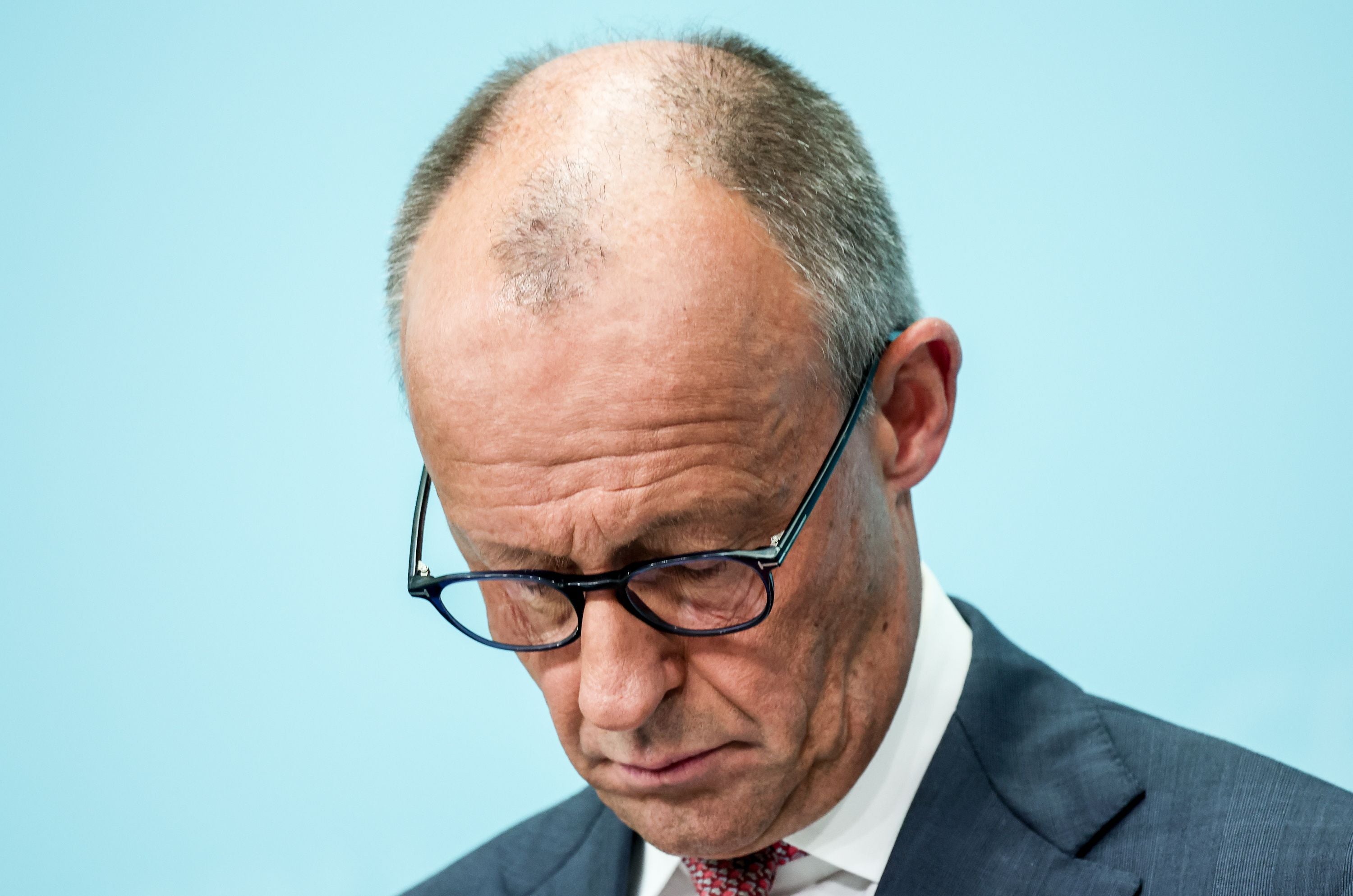 Ein Mann mit Brille und Anzug blickt nach unten vor hellblauem Hintergrund.