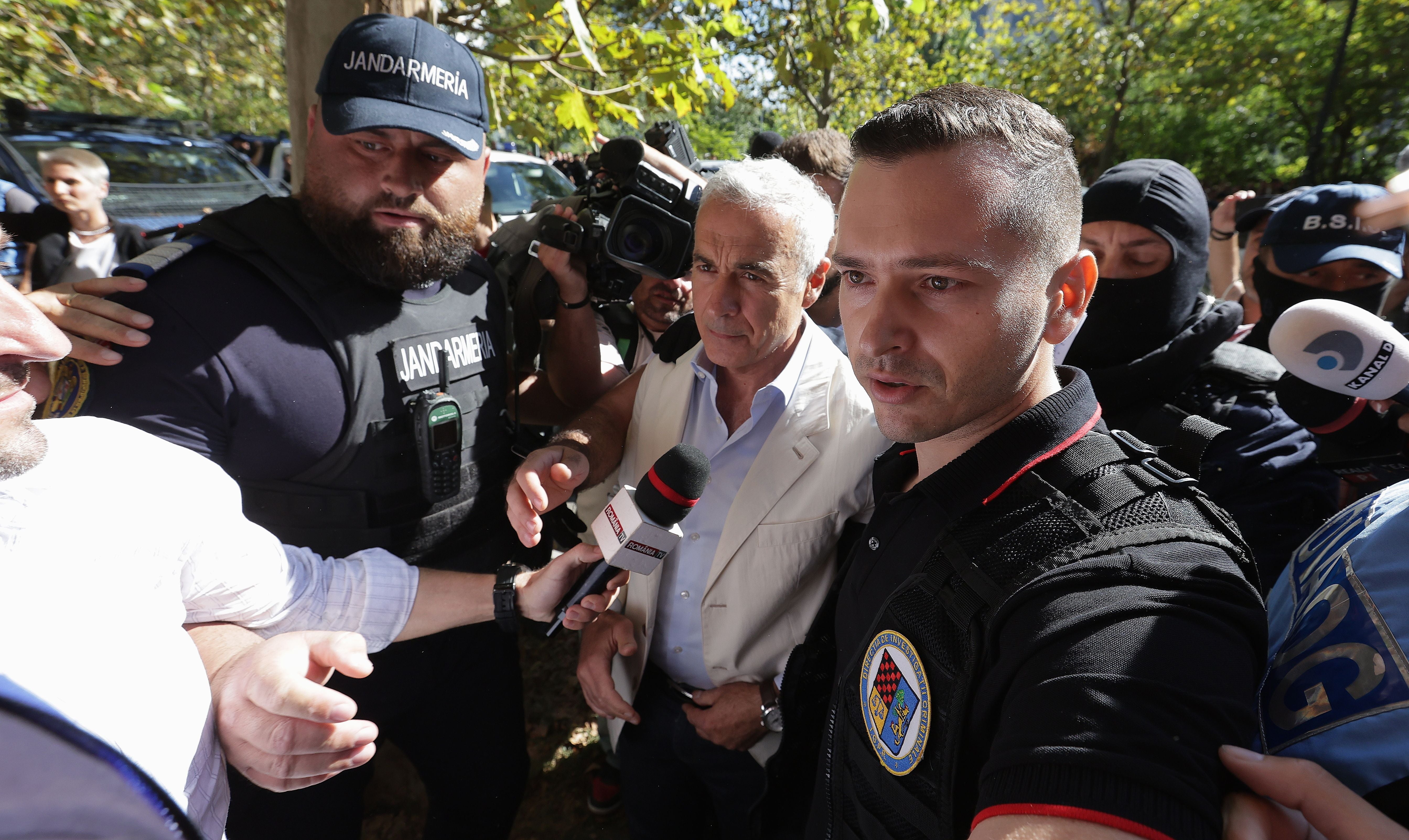 Ein älterer Mann wird von Polizei und Medienvertretern durch eine Menschenmenge begleitet.