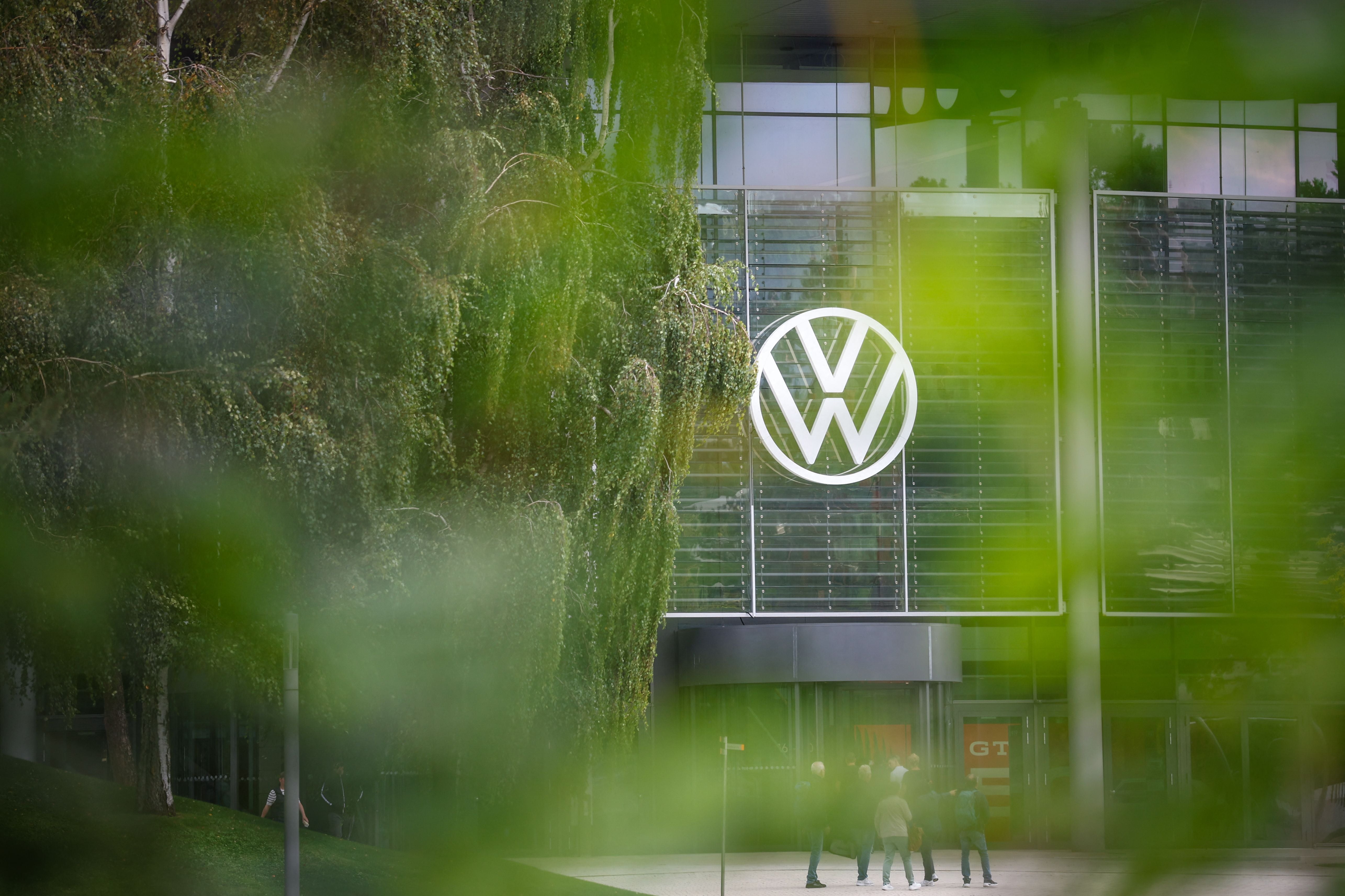 Großes VW-Logo an einem Gebäude mit Glasfront und Bäumen, Menschen stehen davor.