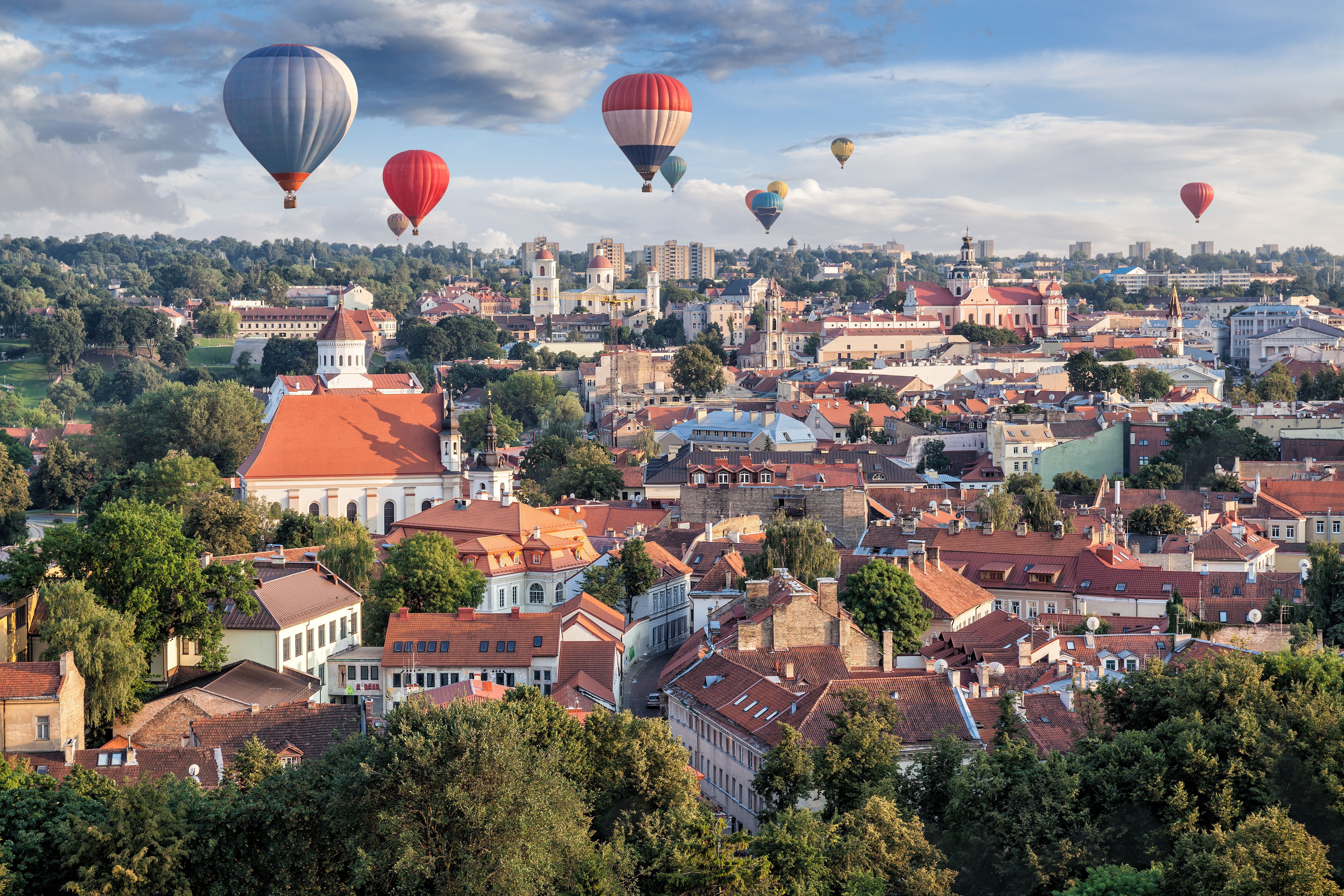 Mehrere Heißluftballons schweben über den Dächern von Vilnius bei Tageslicht.