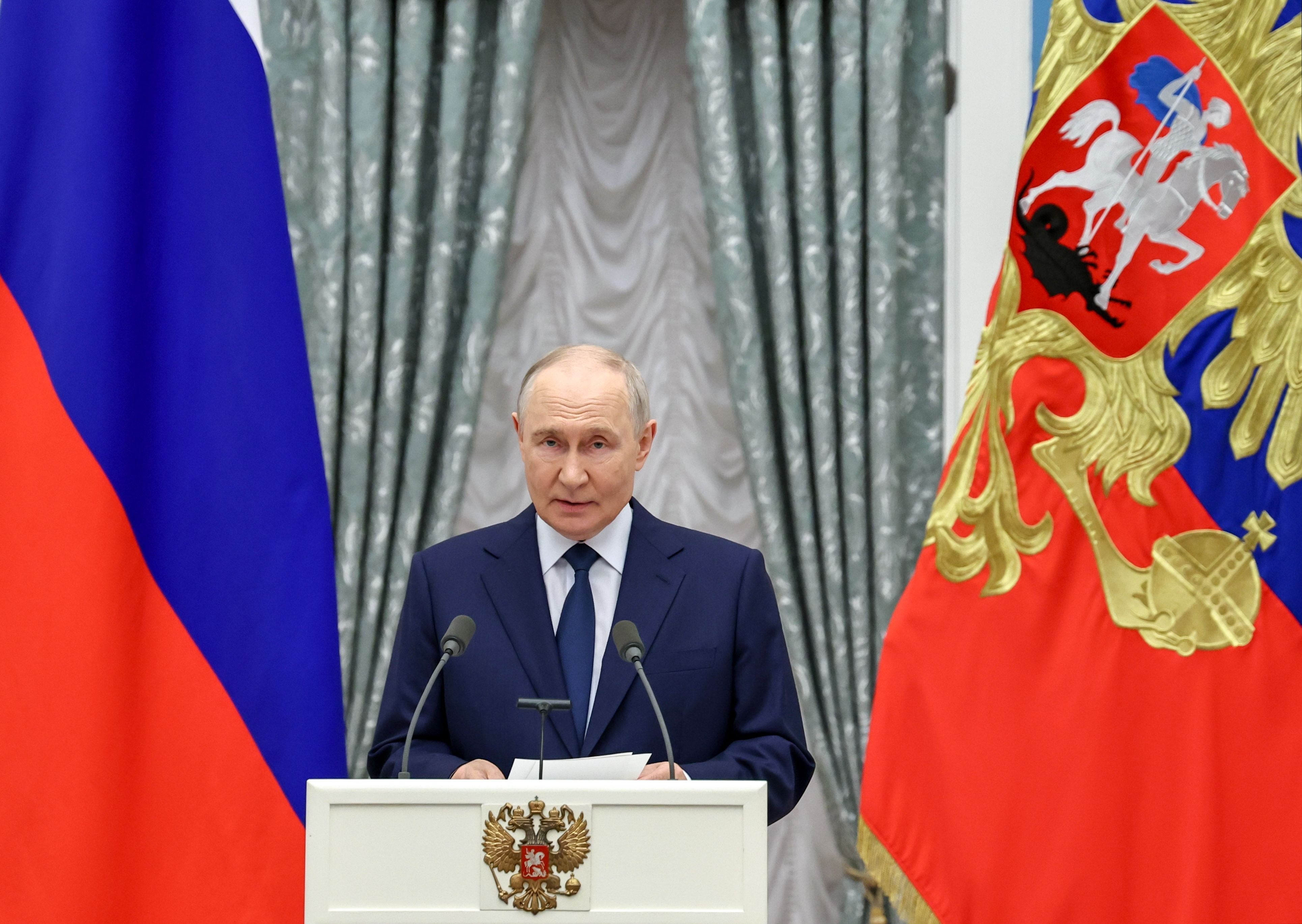 Ein Mann im Anzug spricht an einem Rednerpult zwischen russischen Flaggen und Vorhängen.