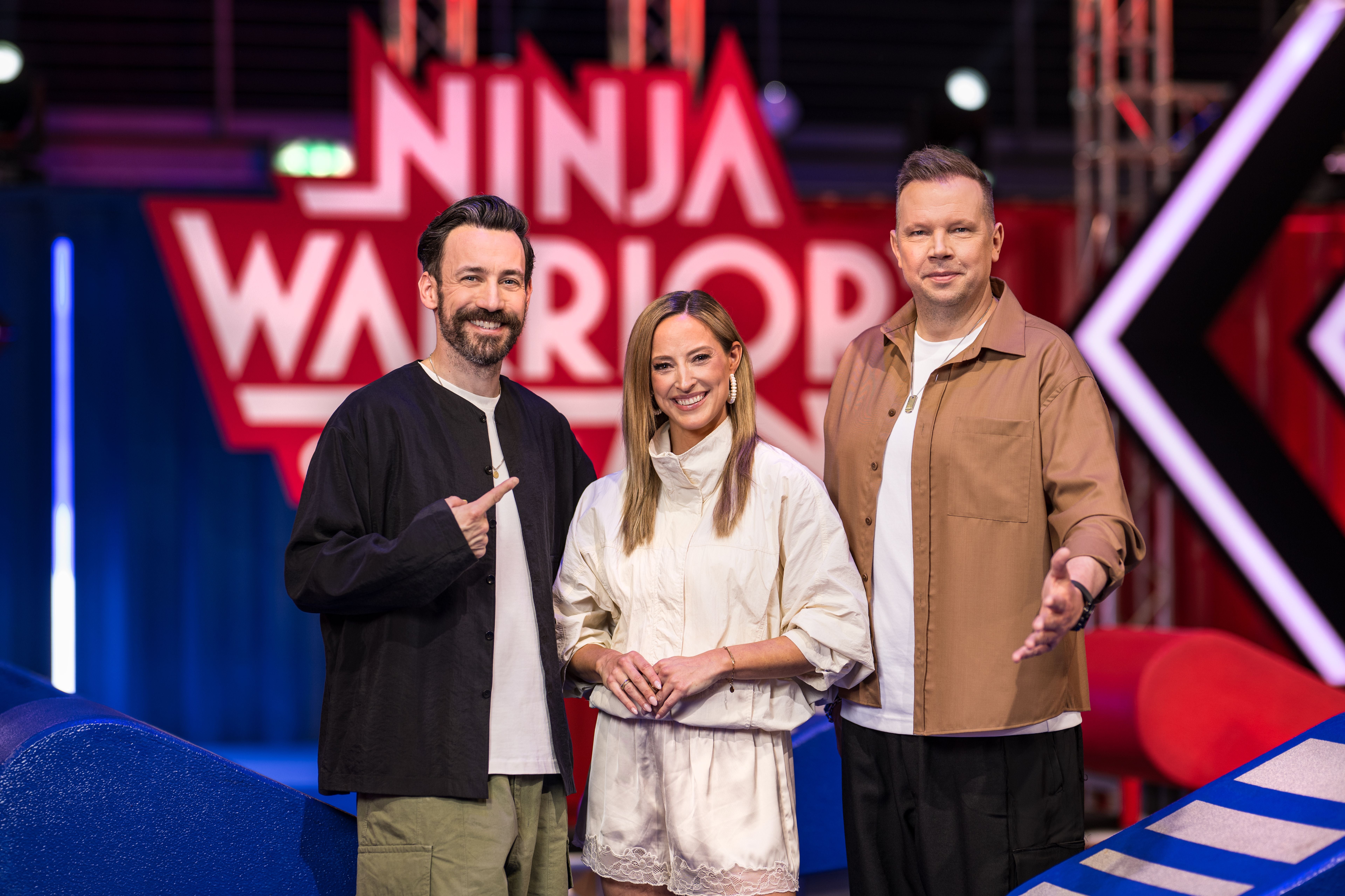 Drei Menschen stehen auf einem „Ninja Warrior“-Set und lächeln in die Kamera.