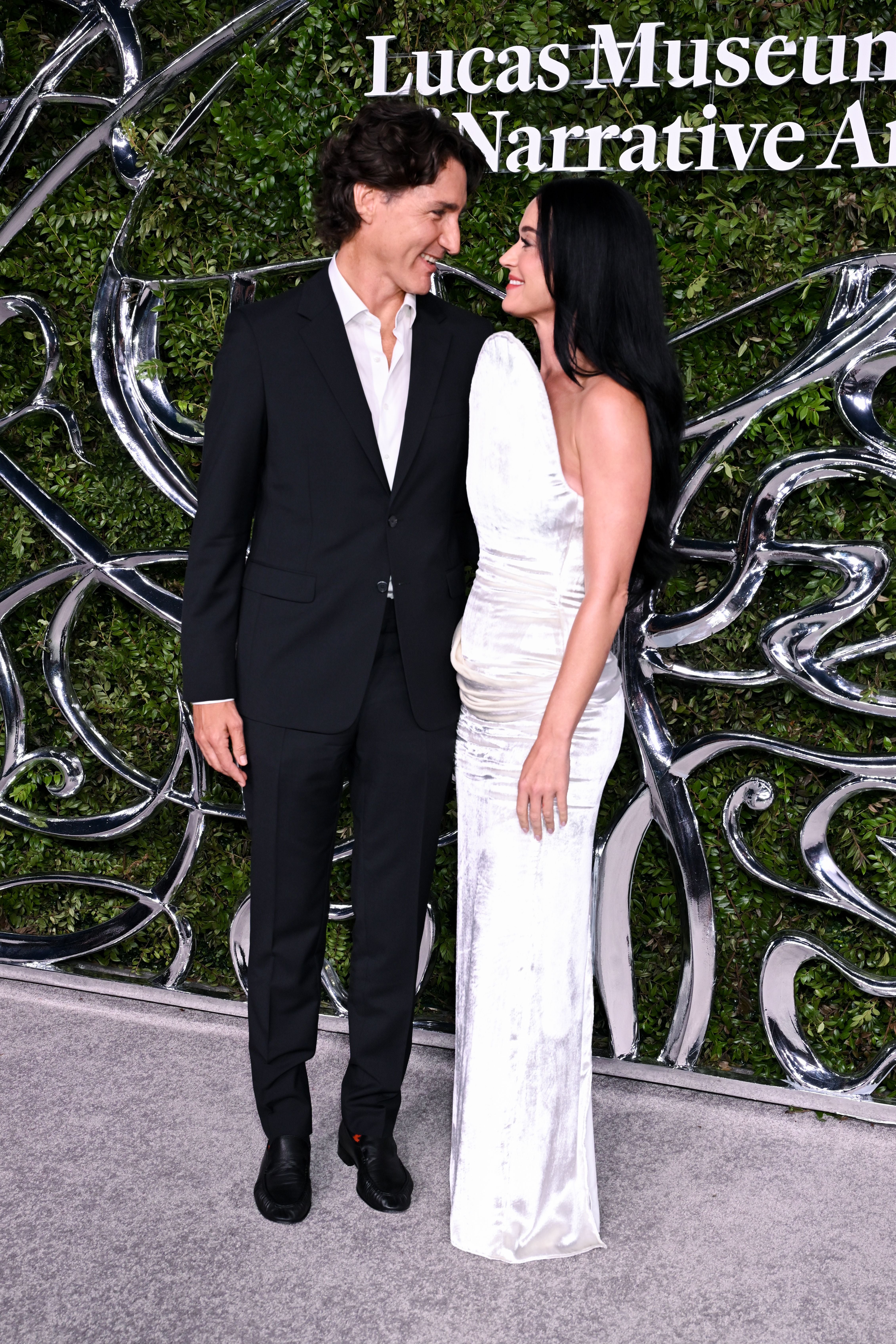 Ein Mann im Anzug und eine Frau im weißen Kleid lächeln sich bei einer Gala vor grünem Hintergrund an.