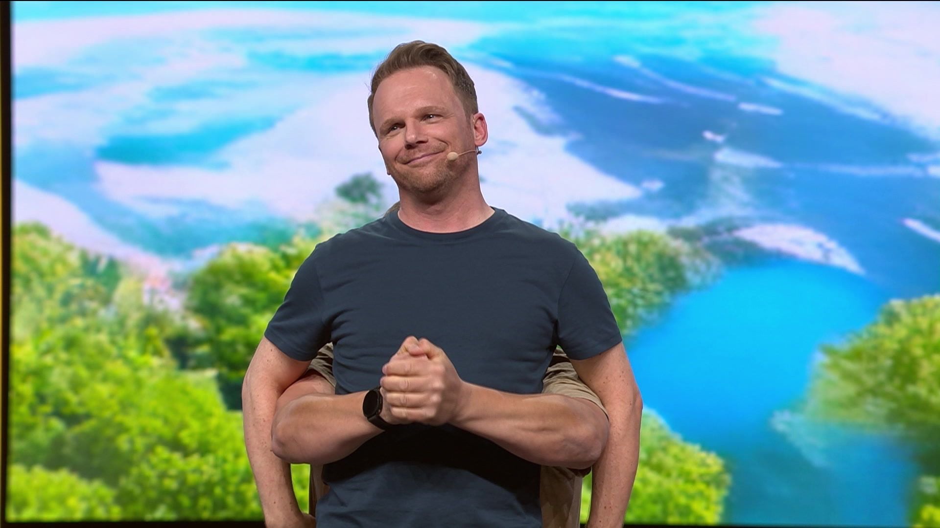 Ein Mann steht mit verschränkten Armen vor einer tropischen Landschaft und trägt ein Mikrofon.