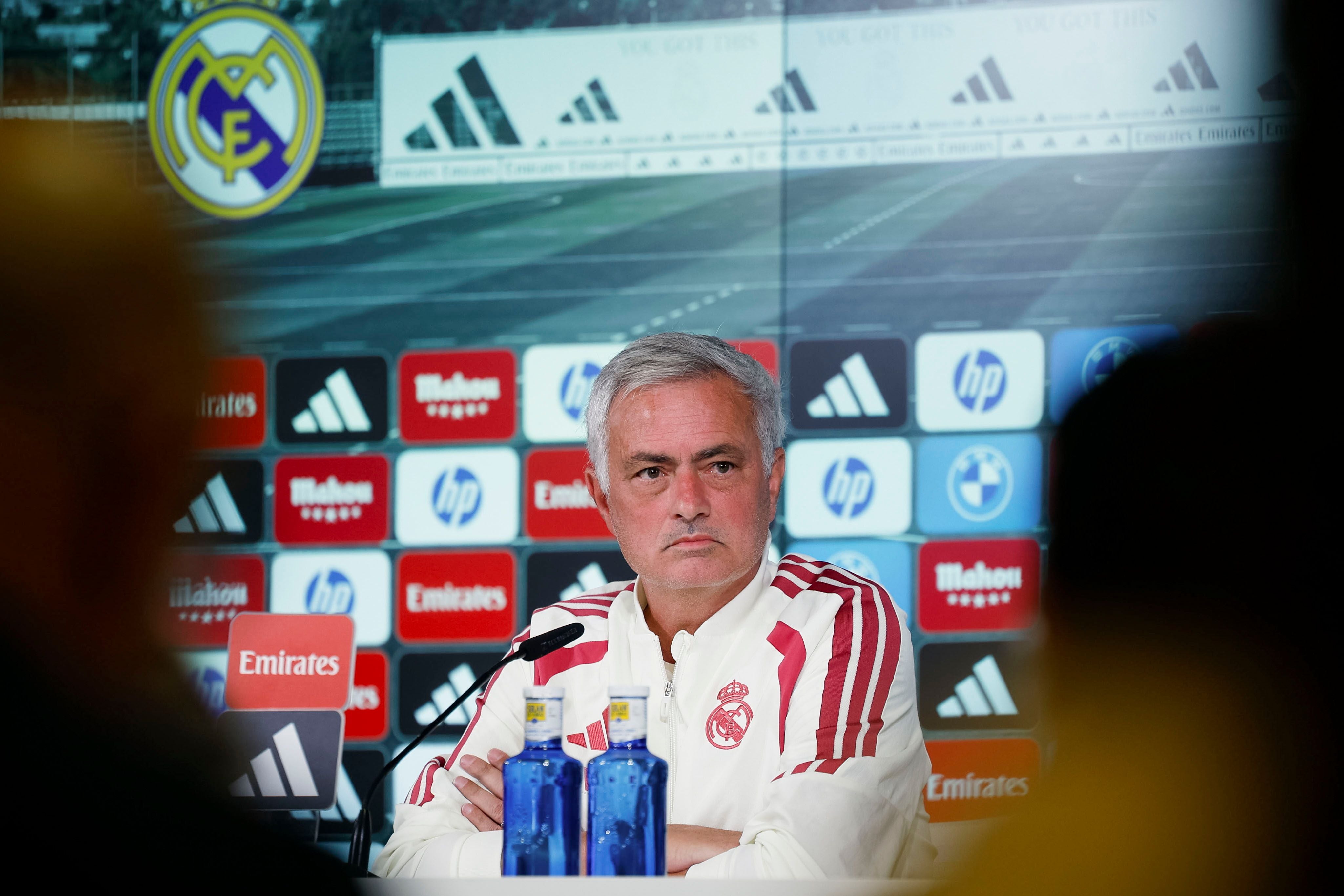 Ein Mann im Real-Madrid-Trainingsanzug spricht auf einer Pressekonferenz mit Mikrofon und Wasserflaschen.