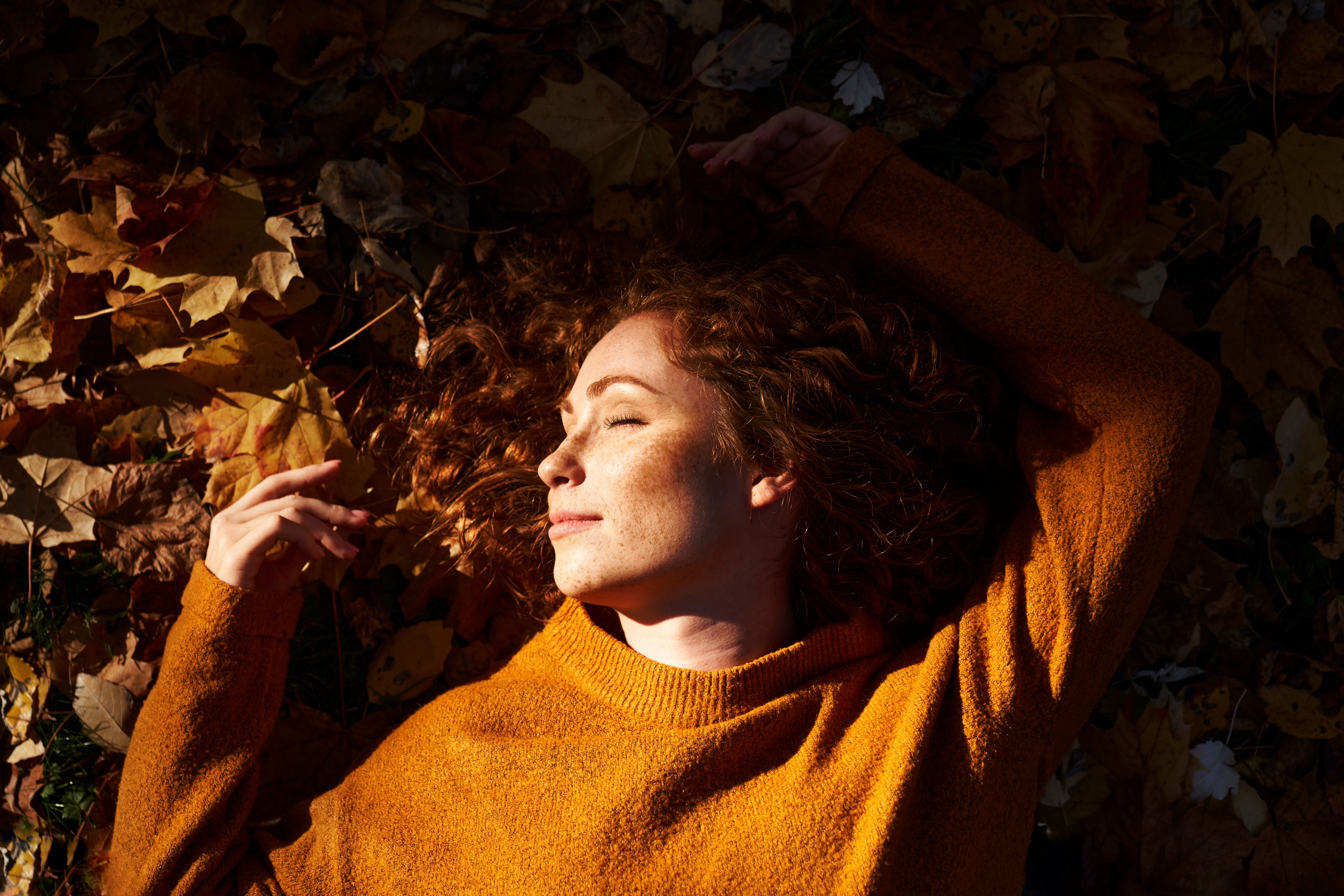 Frau mit lockigem Haar liegt entspannt im Herbstlaub und genießt die Sonne.