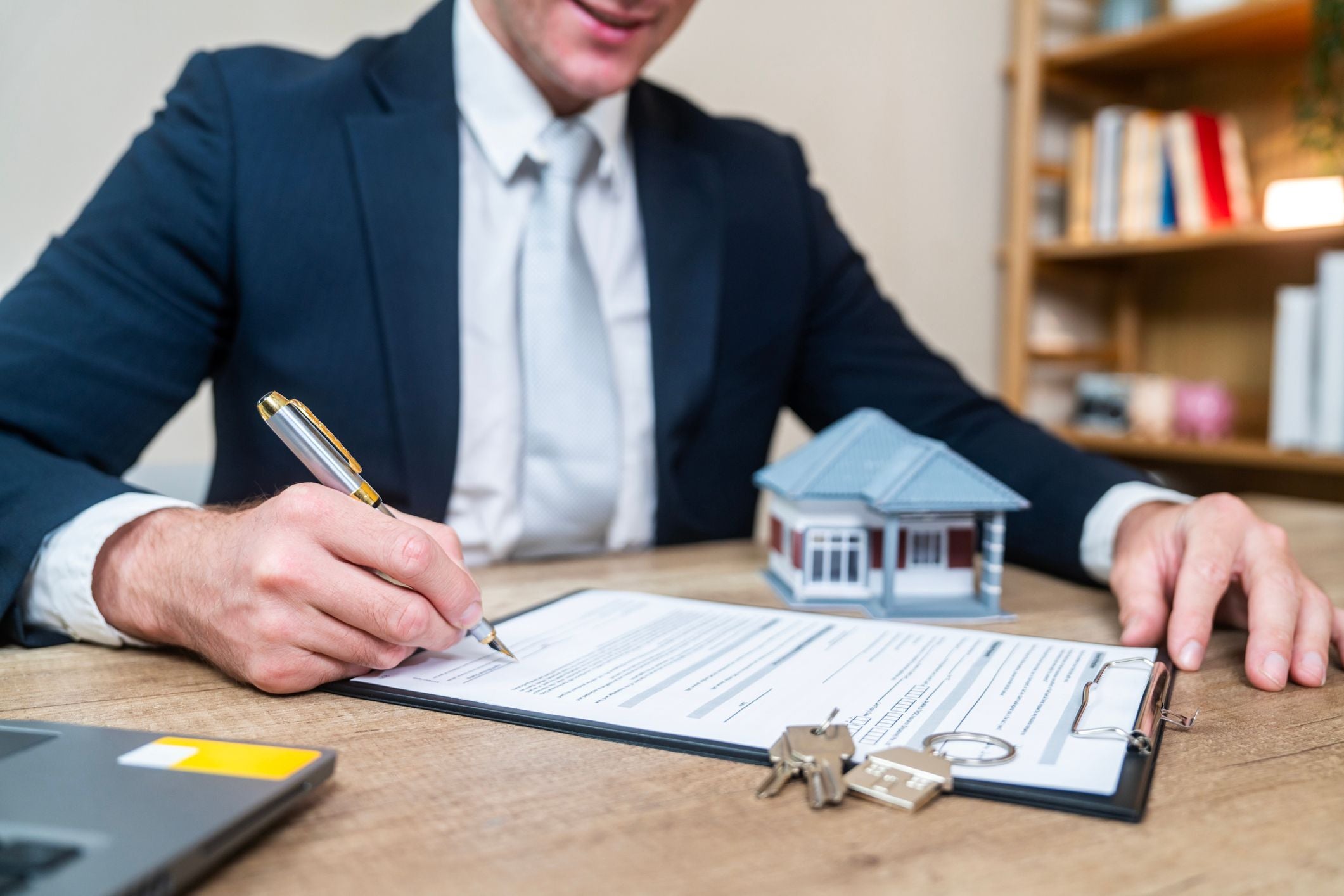 Geschäftsmann unterschreibt Immobilienvertrag, daneben Hausschlüssel und Modellhaus auf dem Tisch.