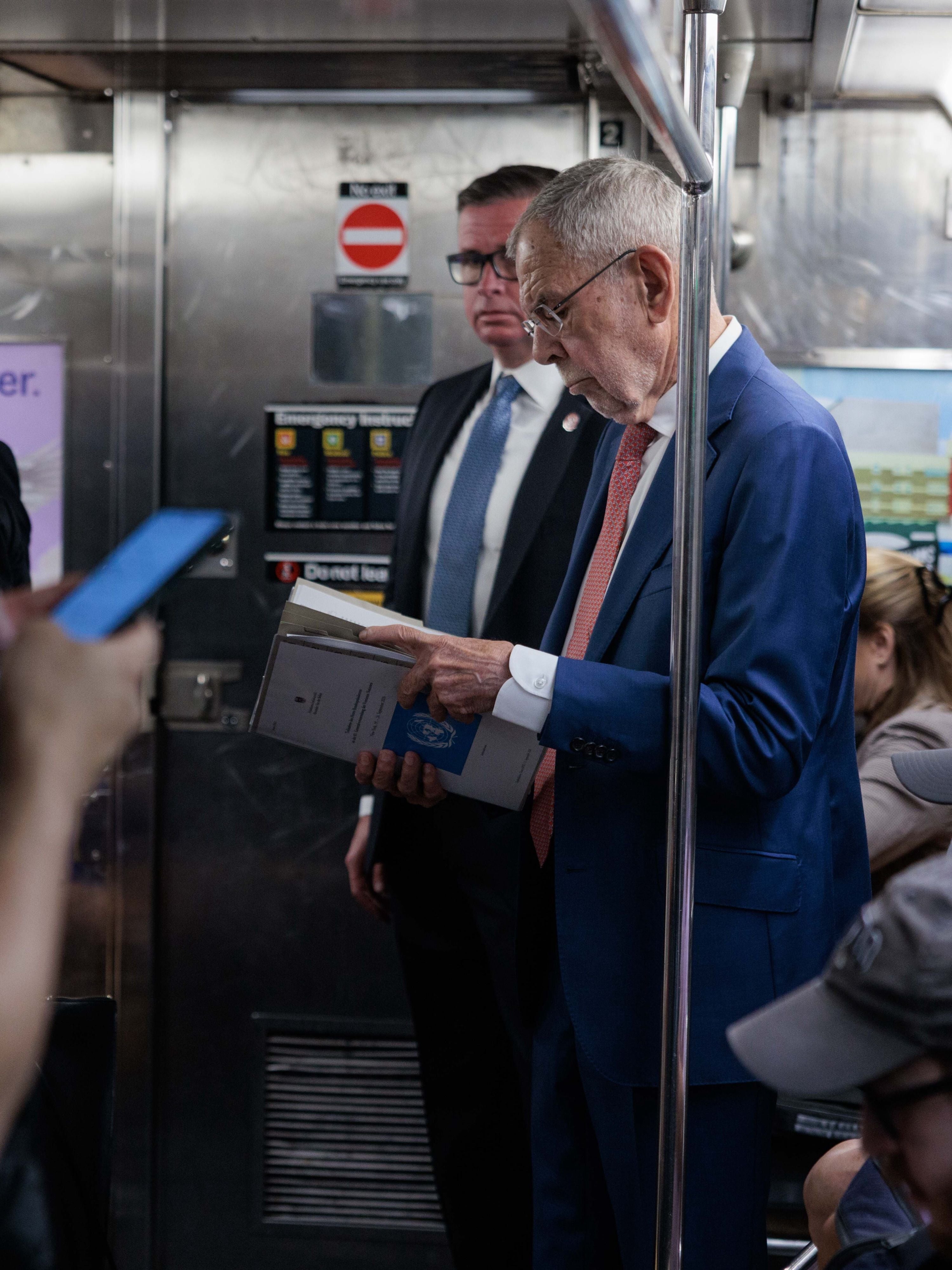 Älterer Mann im Anzug liest Dokumente in einer U-Bahn, ein weiterer Mann steht dahinter.