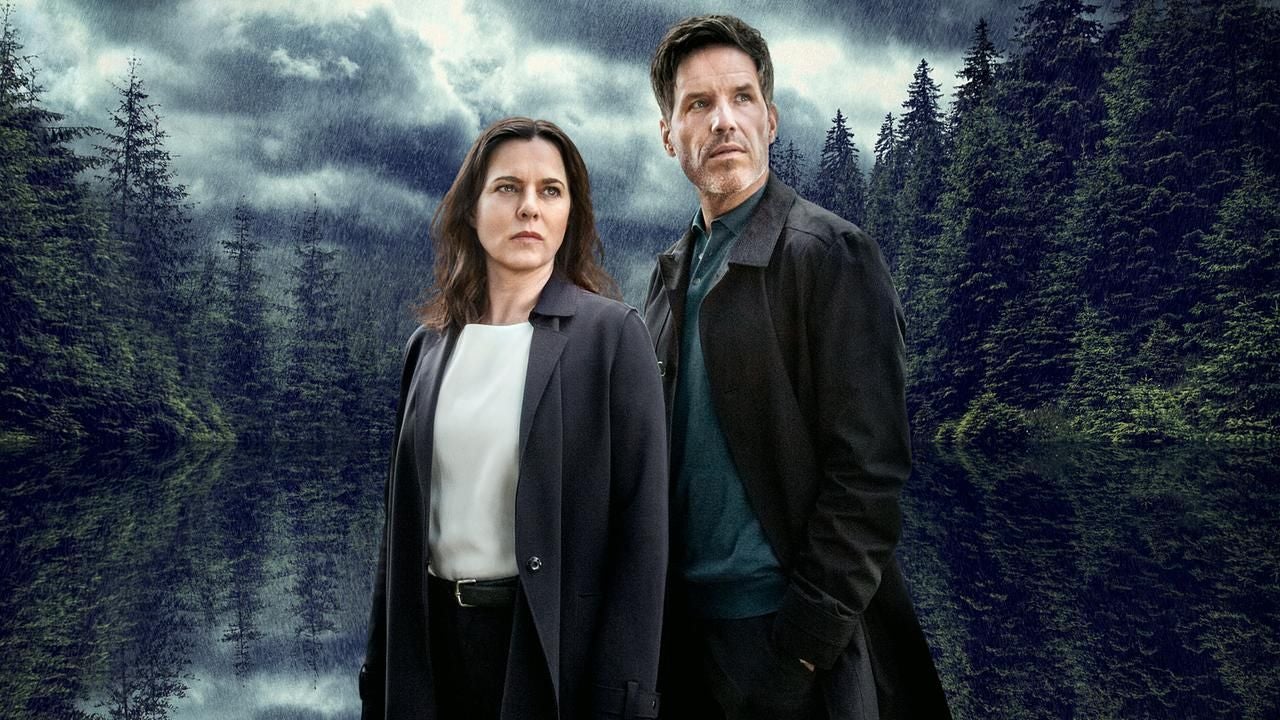 Zwei Personen stehen vor einem dunklen Waldsee bei bewölktem, regnerischem Wetter.