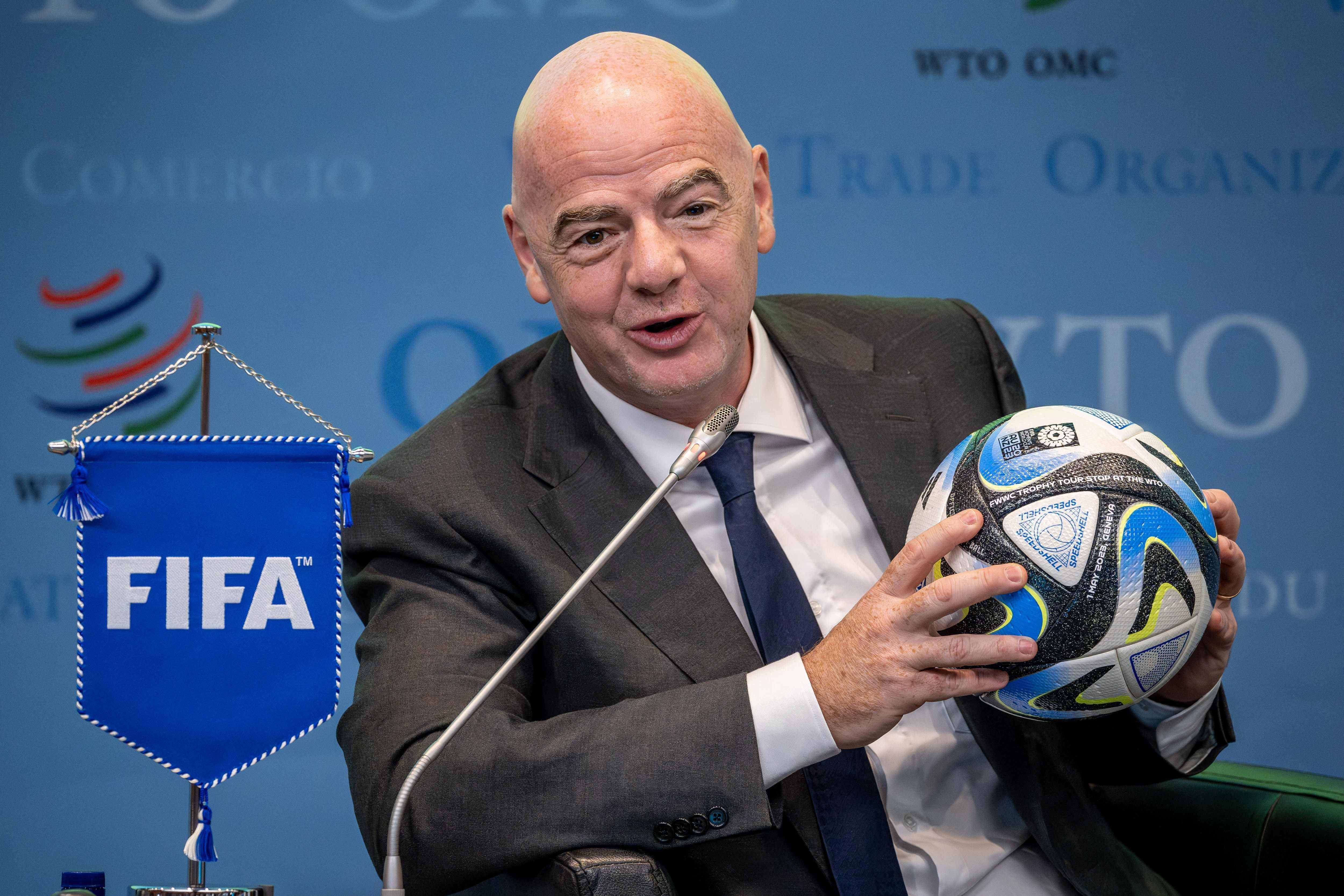 Ein Mann im Anzug hält einen Fußball und sitzt neben einer blauen FIFA-Fahne bei einer Konferenz.