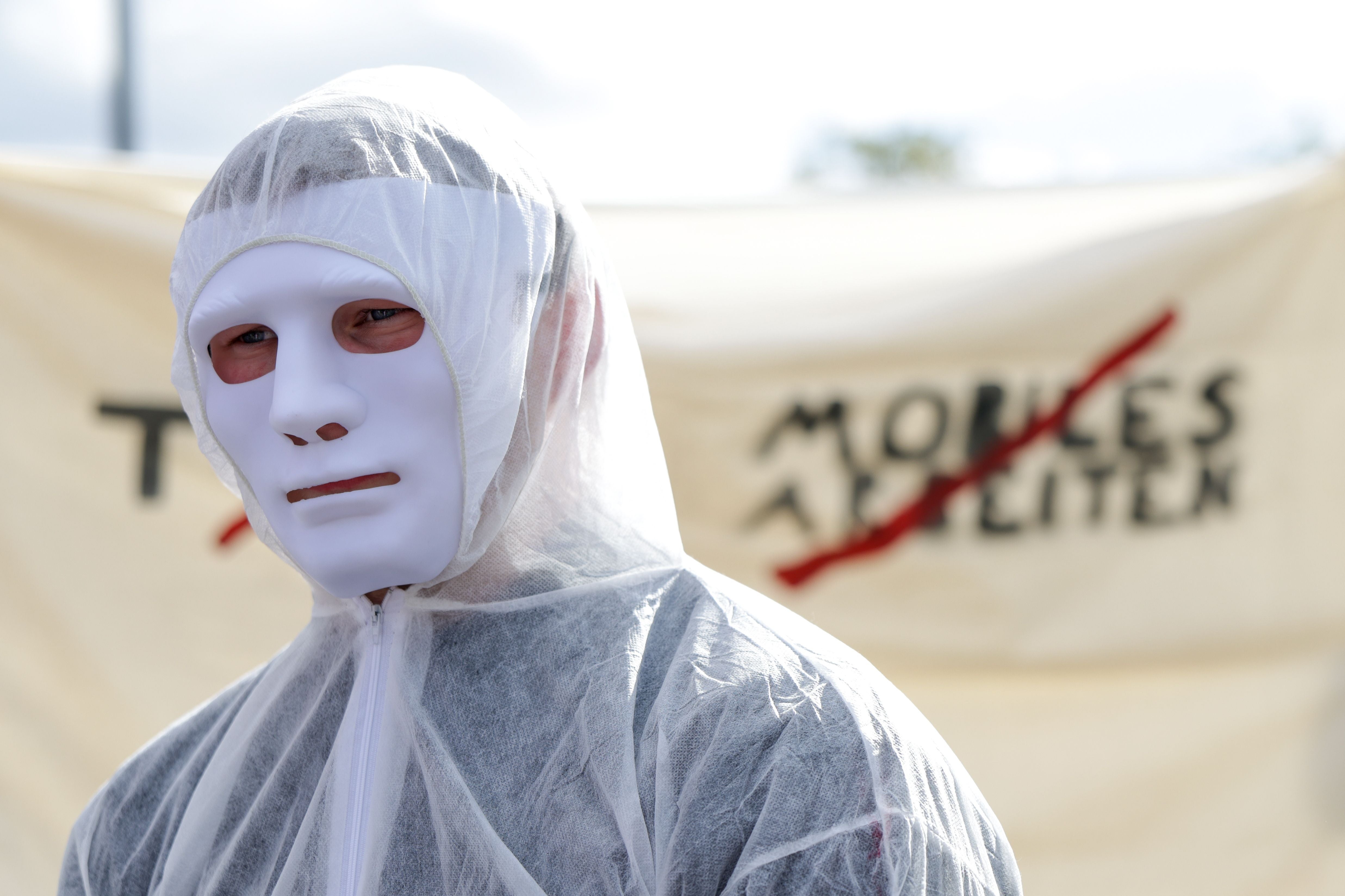 Person mit weißem Overall und weißer Maske bei einer Kundgebung vor einem Transparent.