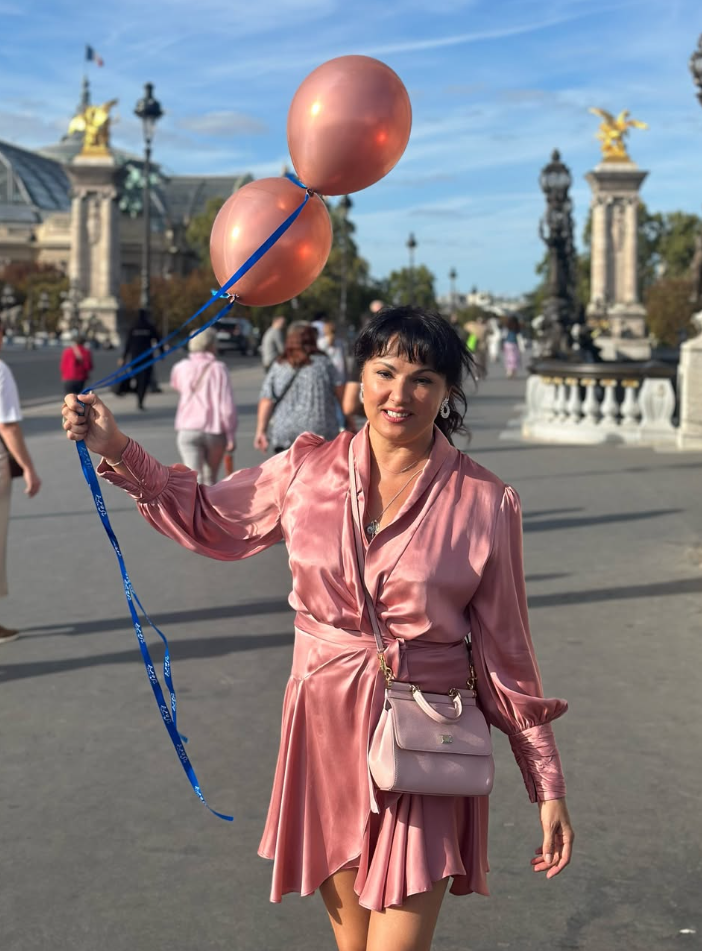 Frau im rosa Kleid hält zwei Ballons und läuft auf einer Pariser Brücke bei Sonnenschein.