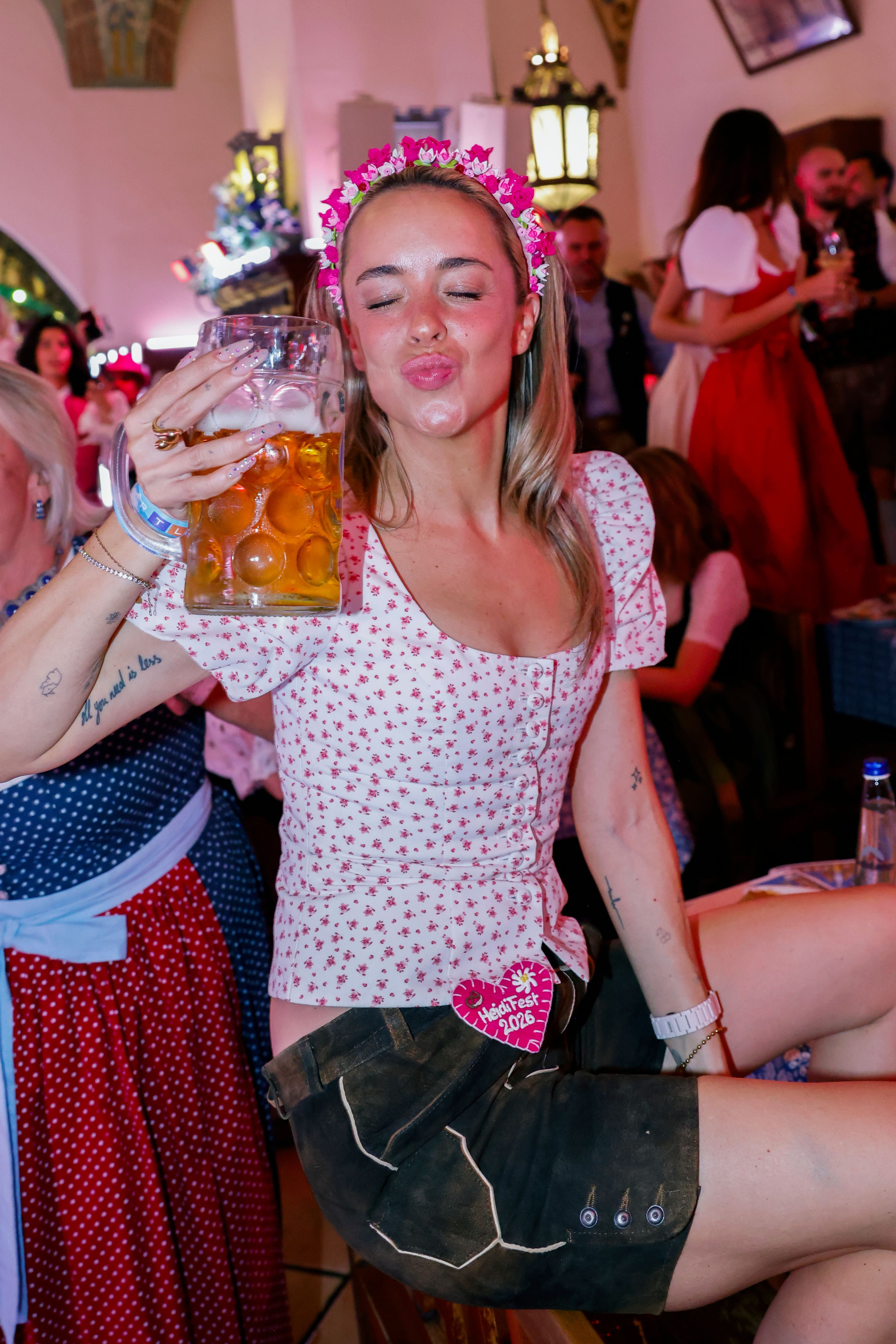 Frau mit Blumenkranz und Bierkrug feiert auf einem Fest im traditionellen Outfit.