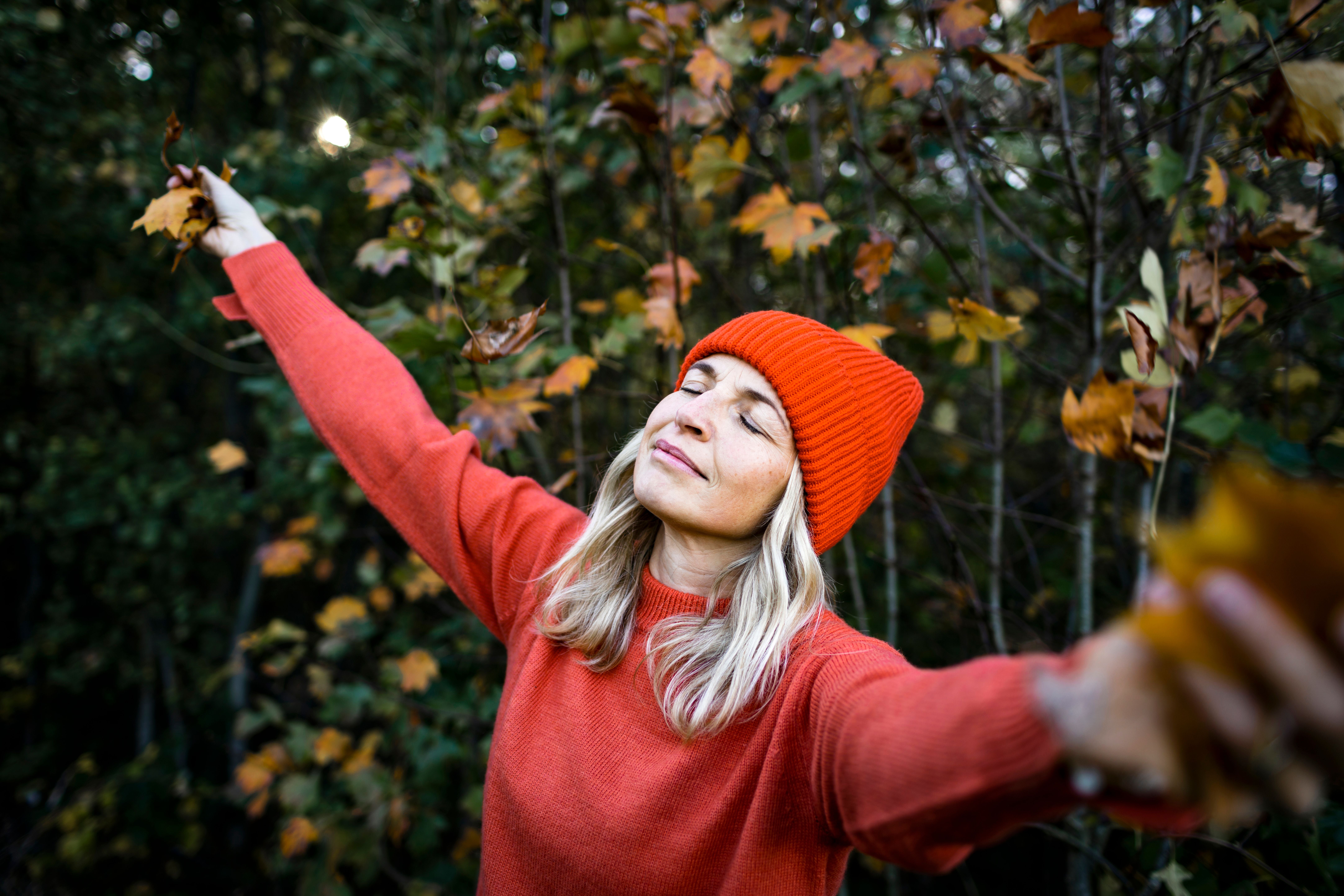 Frau in rotem Pullover und Mütze genießt den Herbst im Wald mit ausgebreiteten Armen.