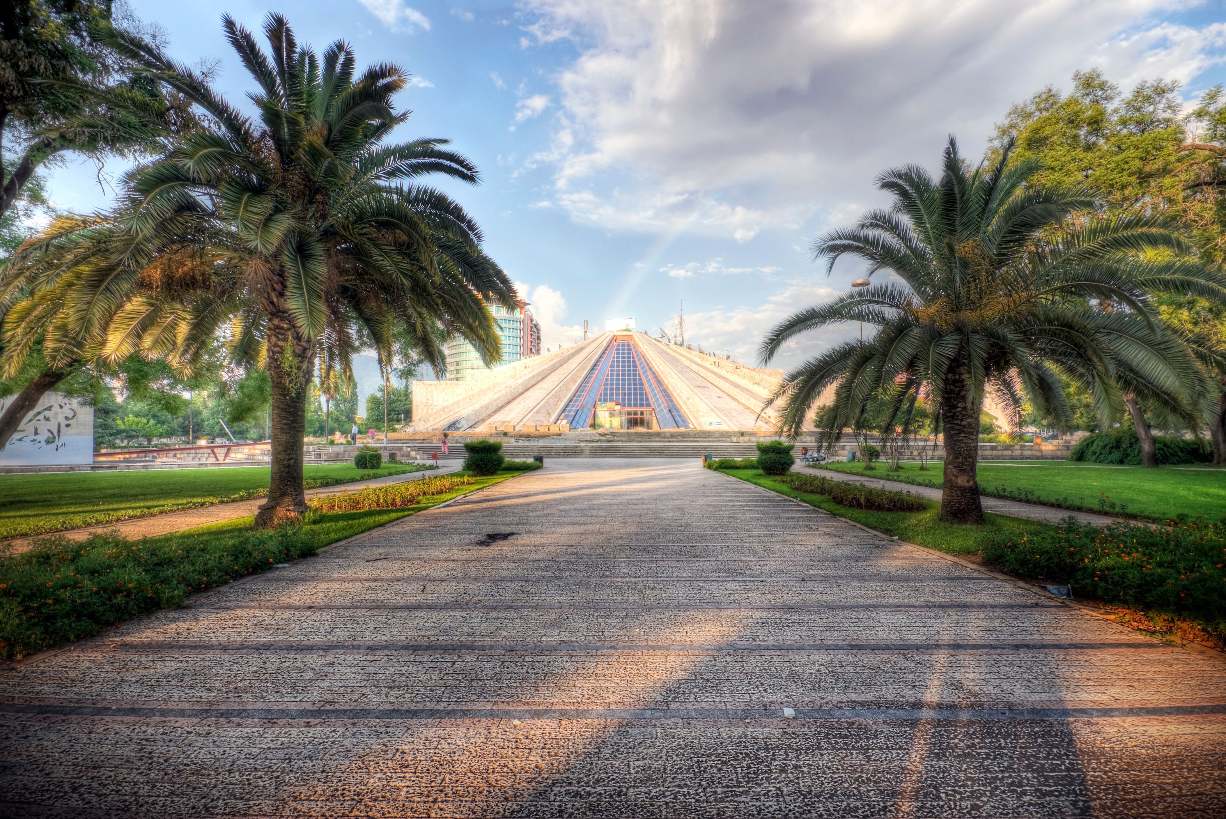 Pyramidenförmiges Gebäude in Tirana, Albanien, mit Palmen und Regenbogen im Hintergrund.
