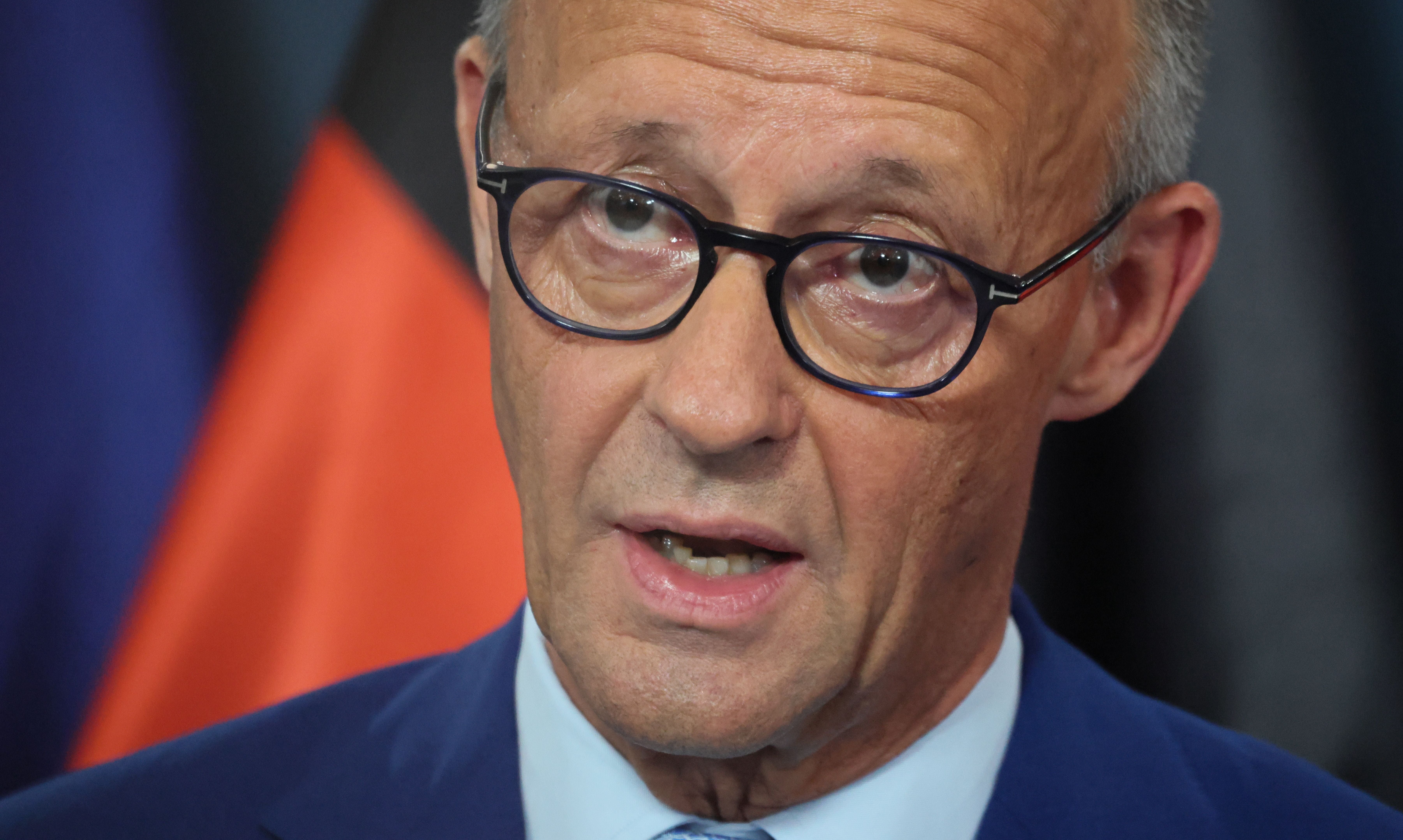 Ein Mann mit Brille spricht, hinter ihm ist unscharf die deutsche Flagge zu sehen.