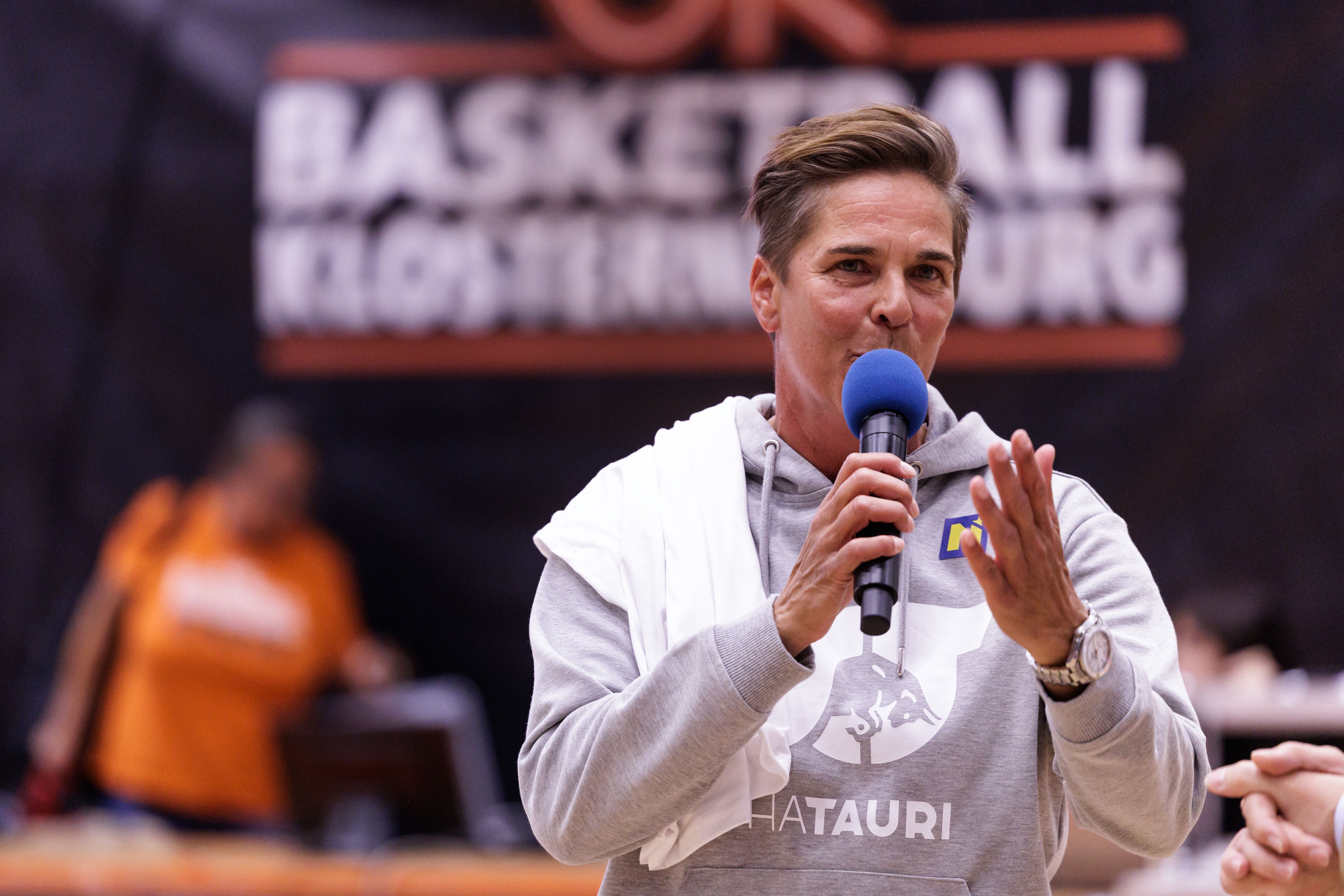 Eine Person spricht mit Mikrofon bei einem Basketball-Event in einer Sporthalle.