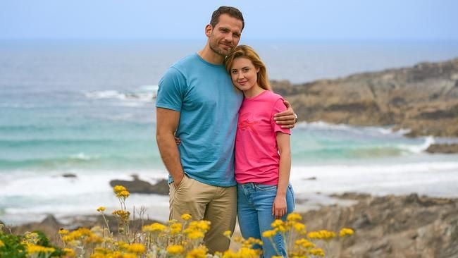 Ein Paar steht umarmt vor einer Küste mit gelben Blumen und Meer im Hintergrund.