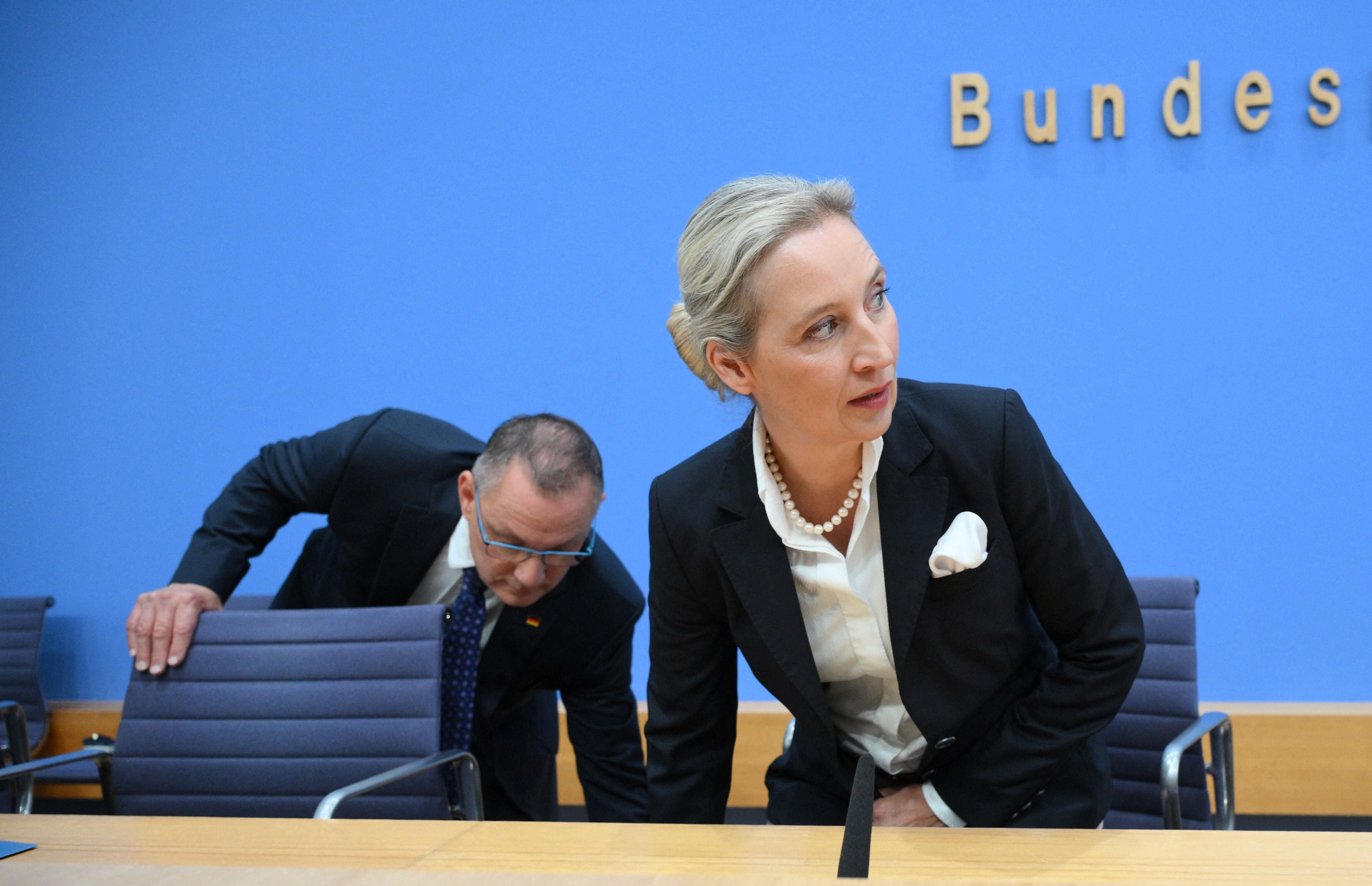 Zwei Personen im Anzug setzen sich bei einer Pressekonferenz vor blaue Wand.