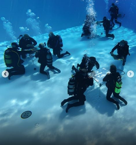 Mehrere Taucher spielen UNO an Tischen auf dem Boden eines Schwimmbeckens unter Wasser.