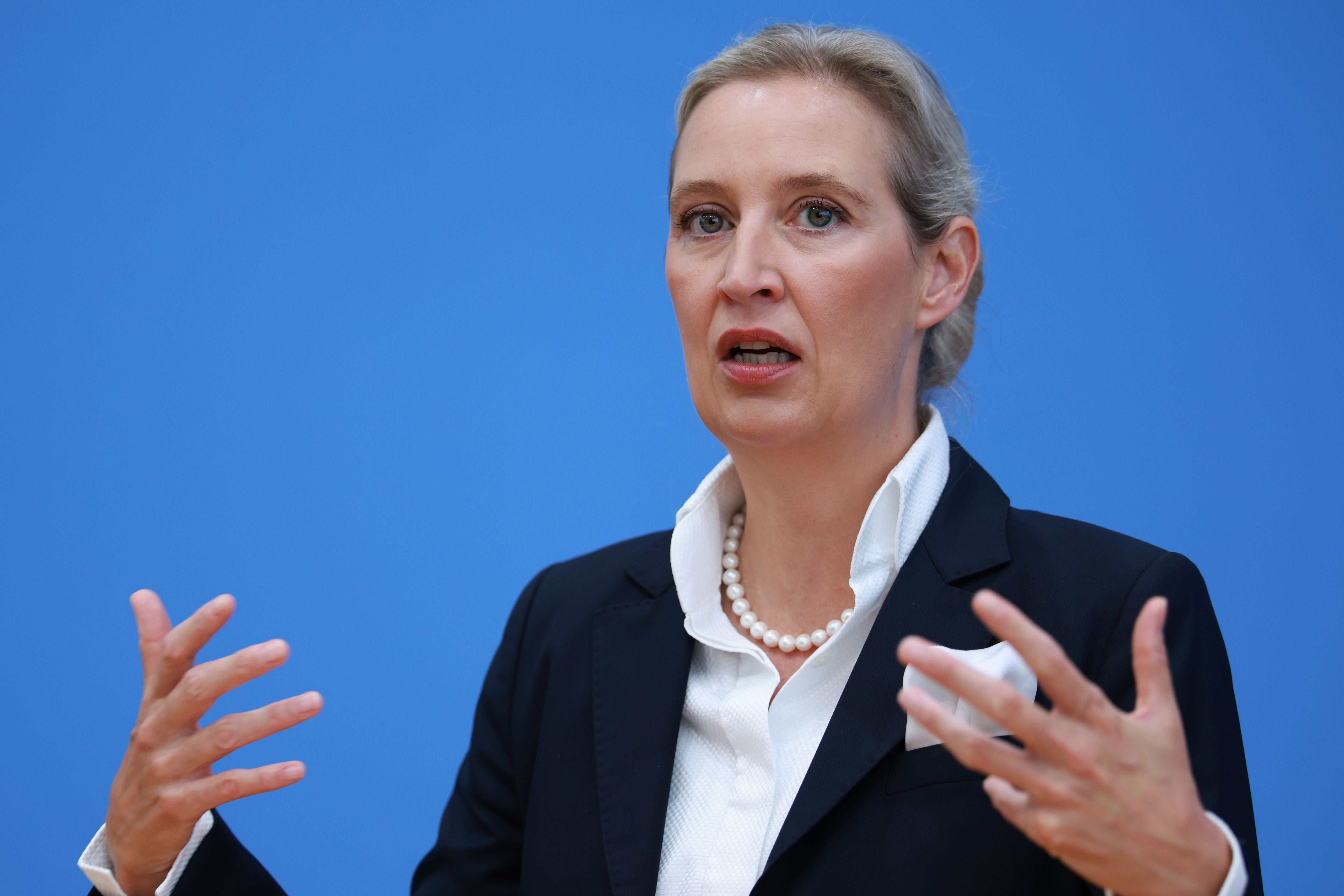 Eine Frau im Anzug spricht gestikulierend vor blauem Hintergrund bei einer Pressekonferenz.