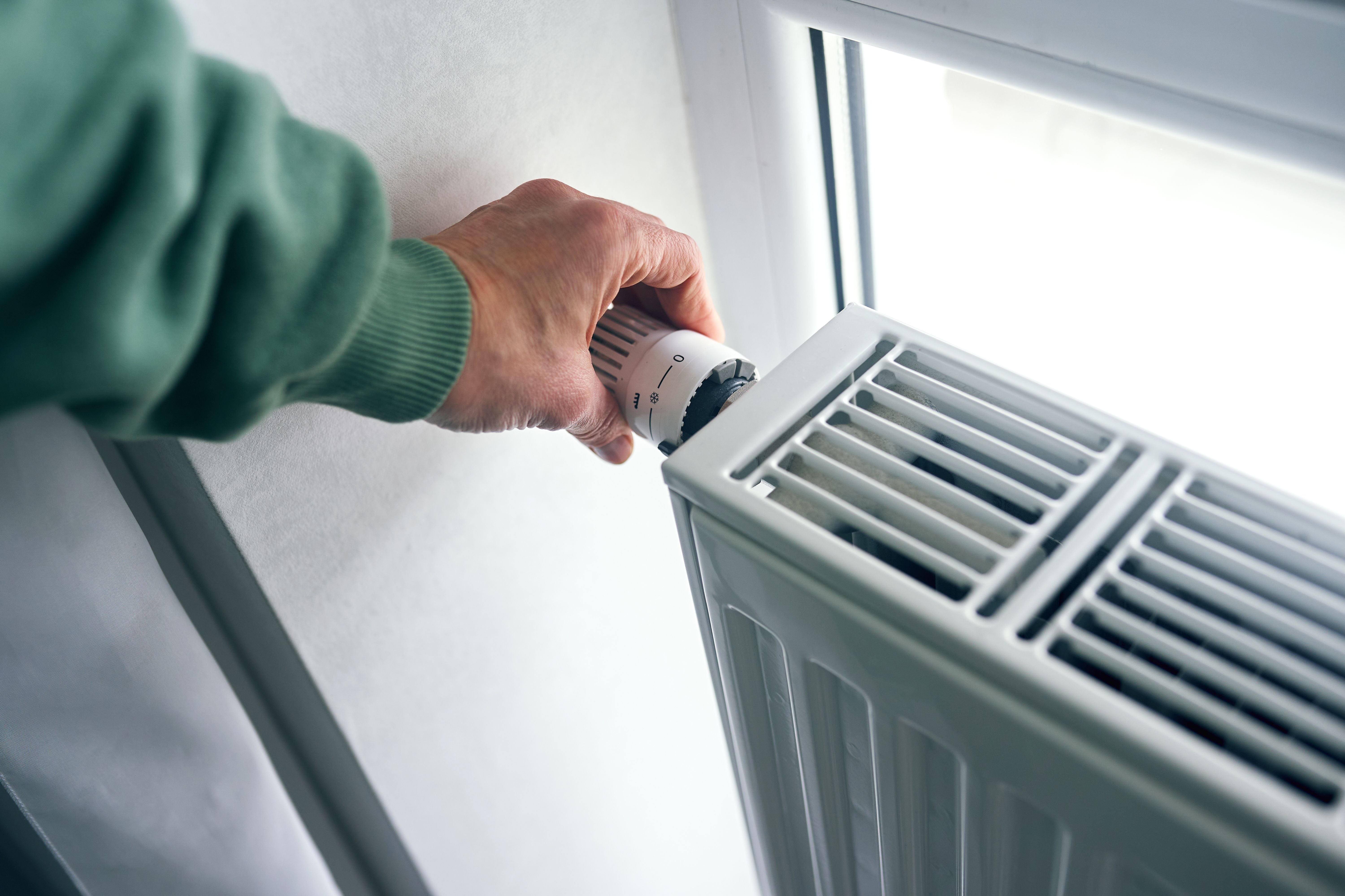 Hand dreht am Thermostat eines Heizkörpers neben einem Fenster, um die Temperatur zu regeln.