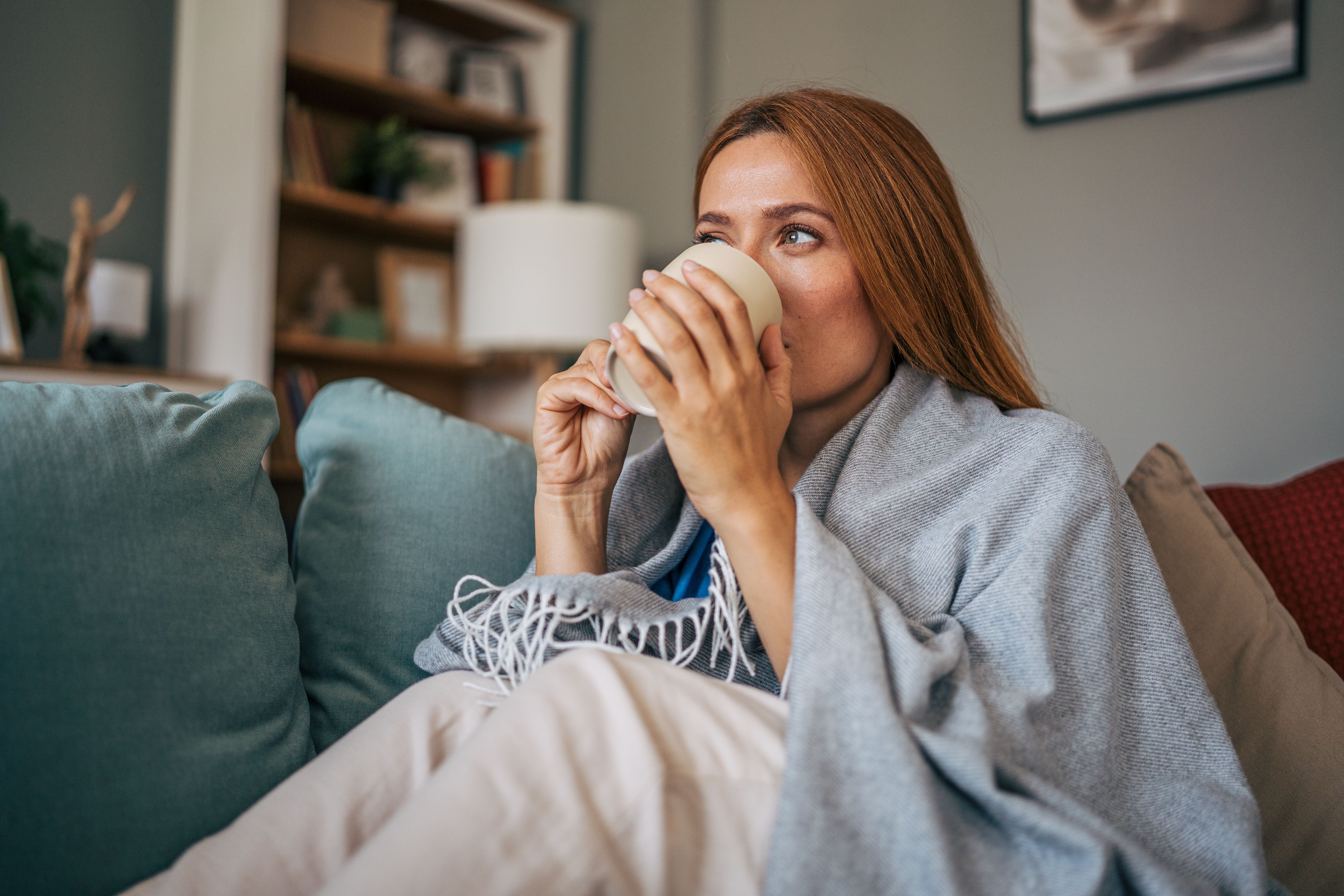 Frau sitzt mit Decke auf dem Sofa und trinkt aus einer Tasse, wirkt entspannt und gemütlich.