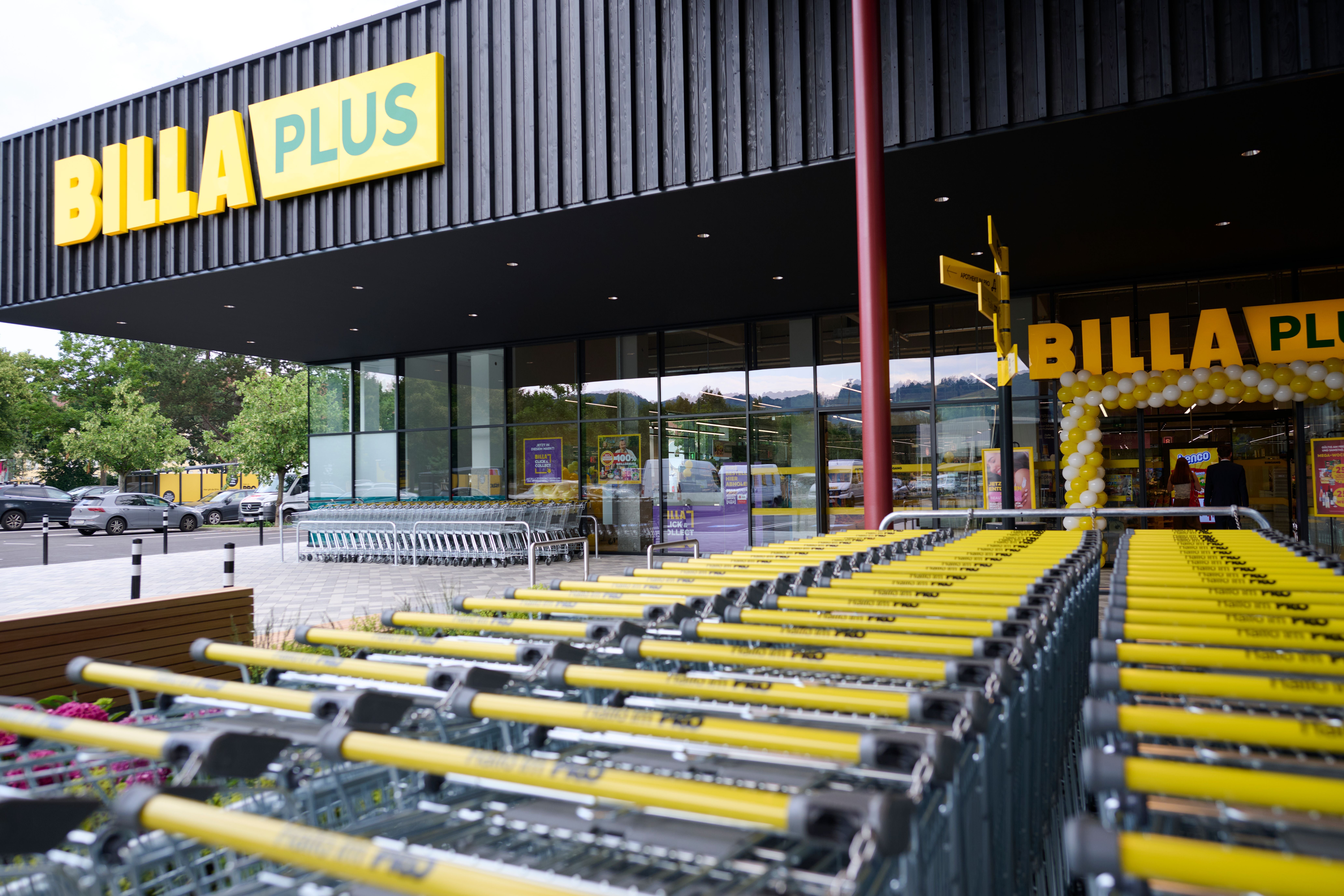 Einkaufswagen mit gelben Griffen vor dem Eingang eines Billa Plus Supermarkts.