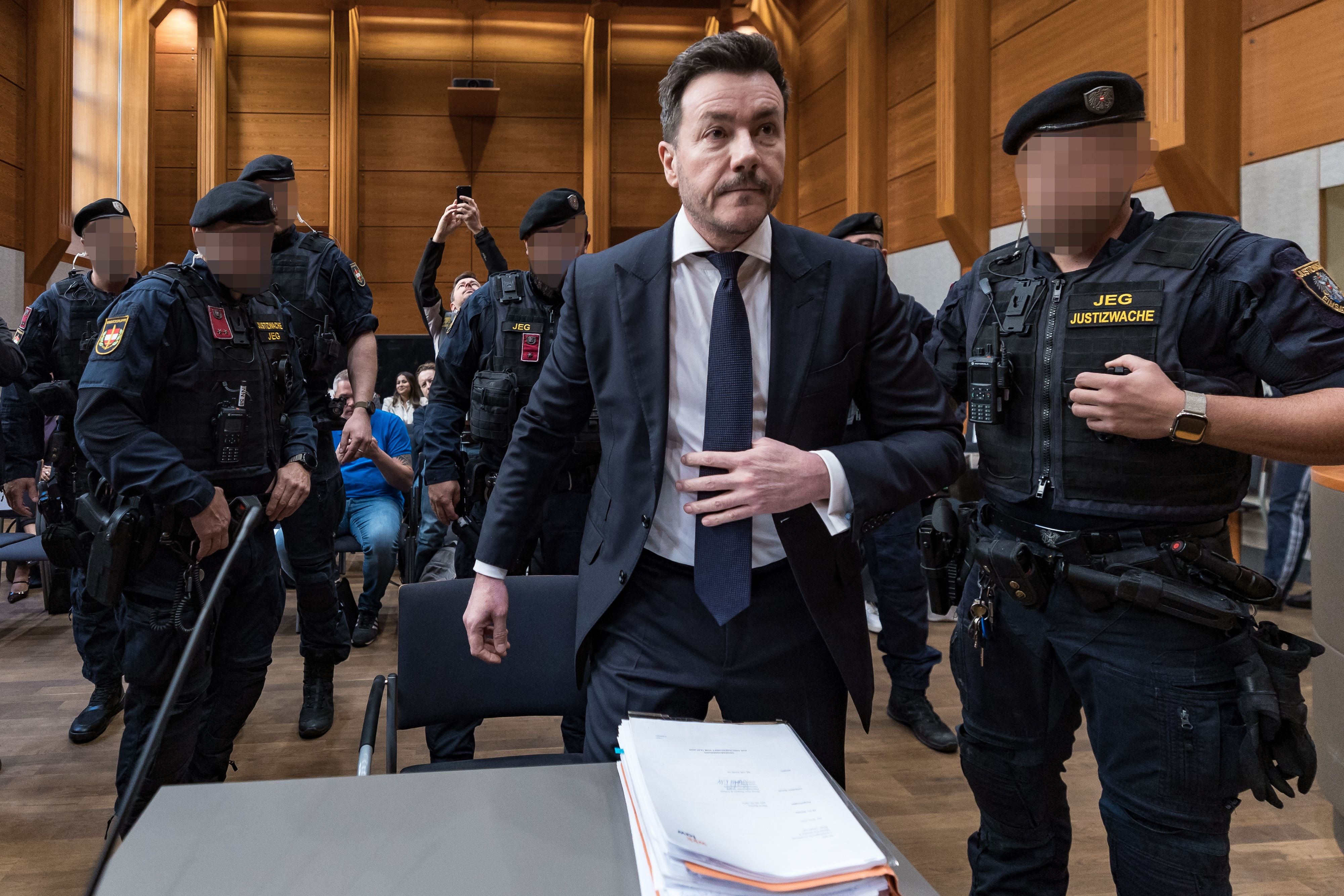 Ein Mann im Anzug steht im Gerichtssaal, umgeben von Justizwache-Beamten in Uniform.