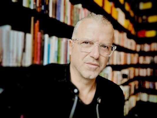 Ein Mann mit Brille sitzt vor einem Bücherregal in einer Bibliothek oder Buchhandlung.