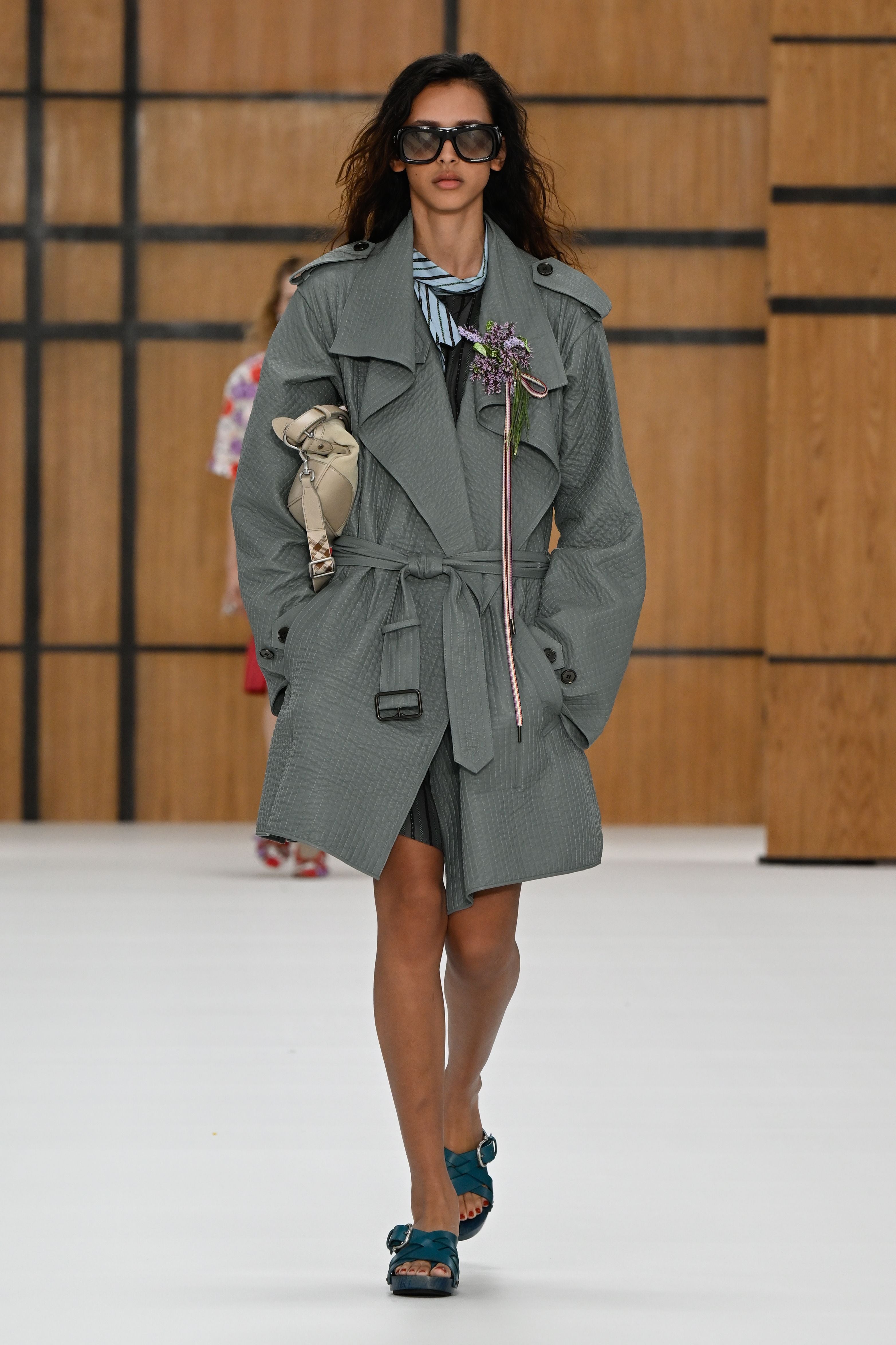 Model trägt grauen Trenchcoat, blaue Sandalen und Sonnenbrille auf dem Laufsteg.