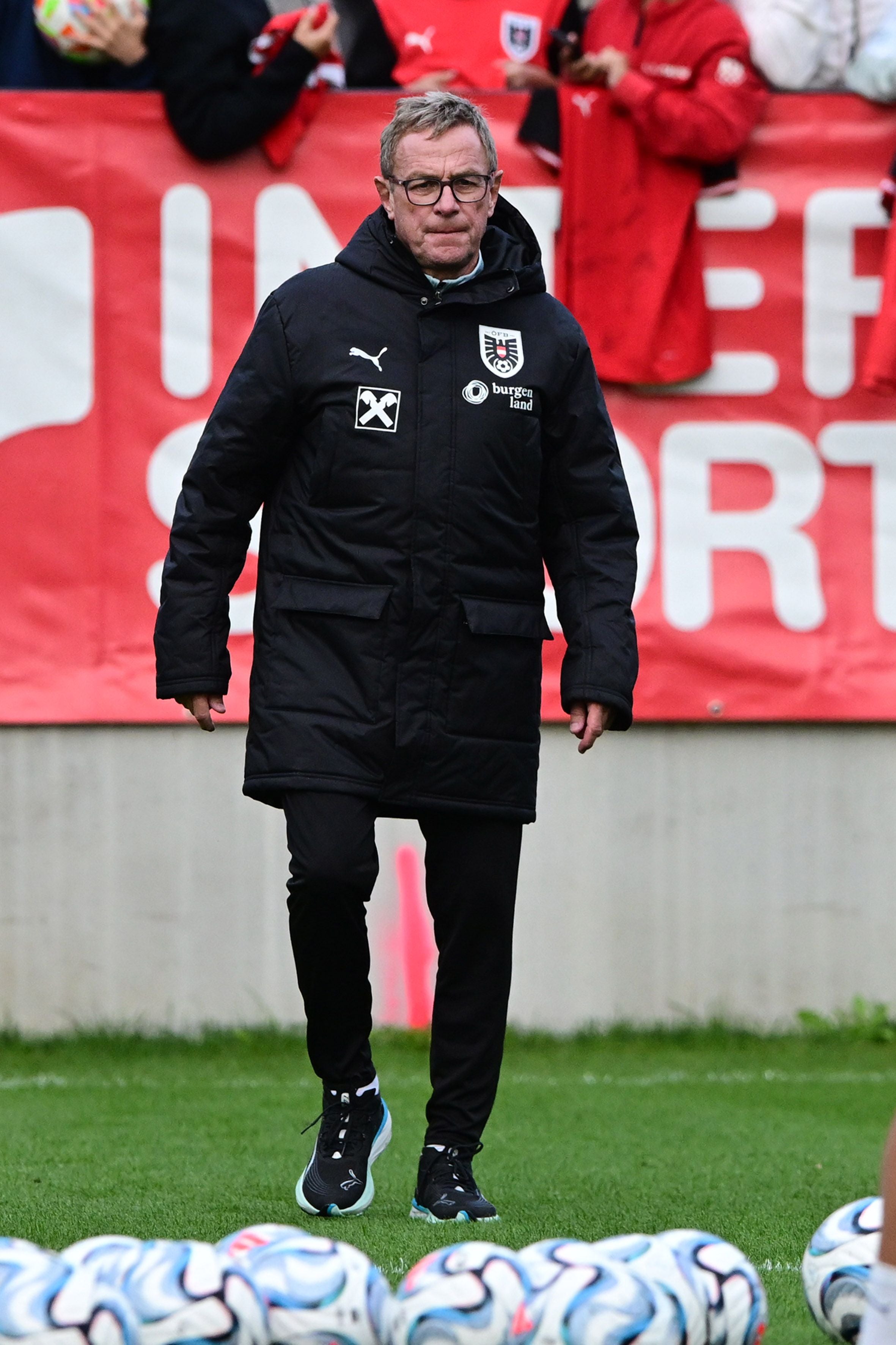 Ein Mann im schwarzen Mantel beim Fußballtraining, im Hintergrund Fans und mehrere Fußbälle.