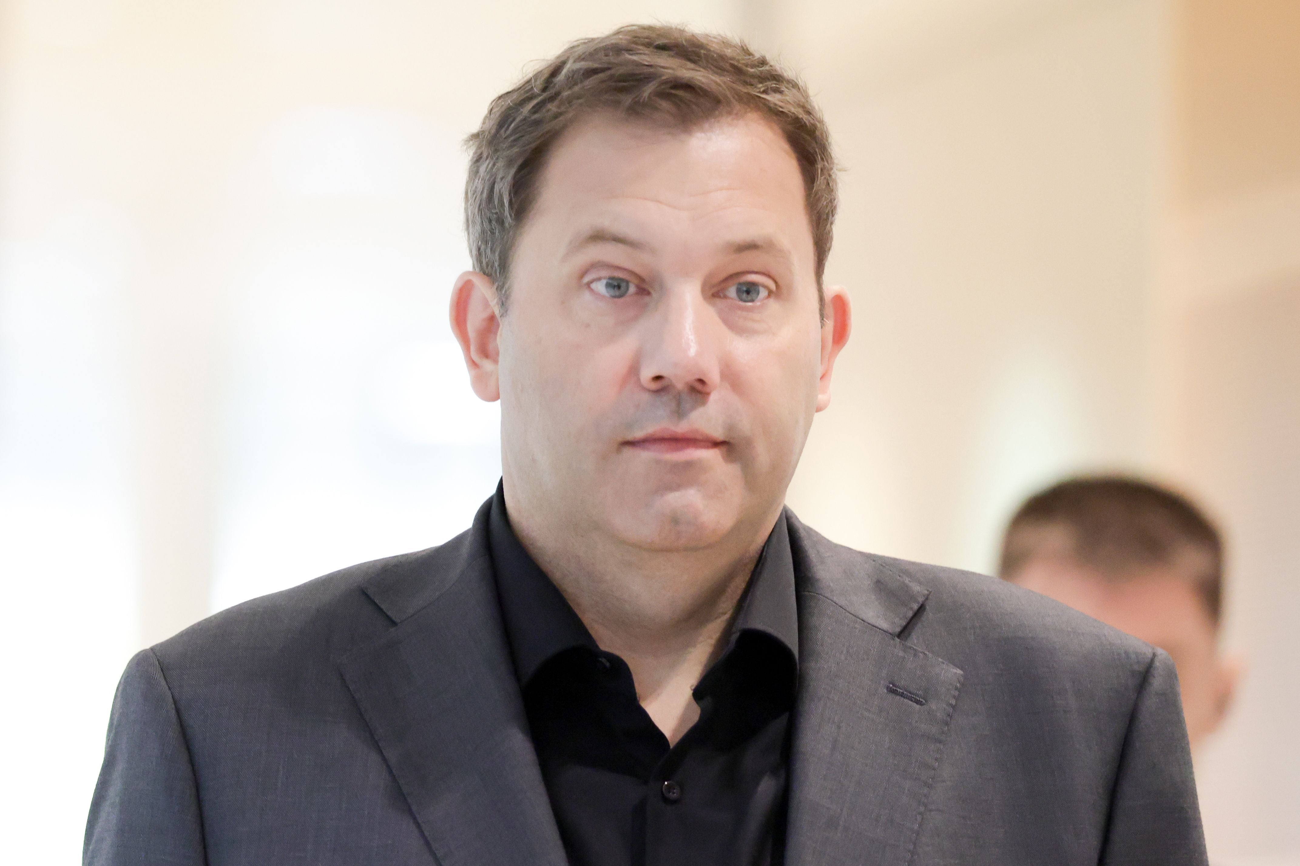 Ein Mann im grauen Anzug blickt ernst in die Kamera, unscharfer Hintergrund.
