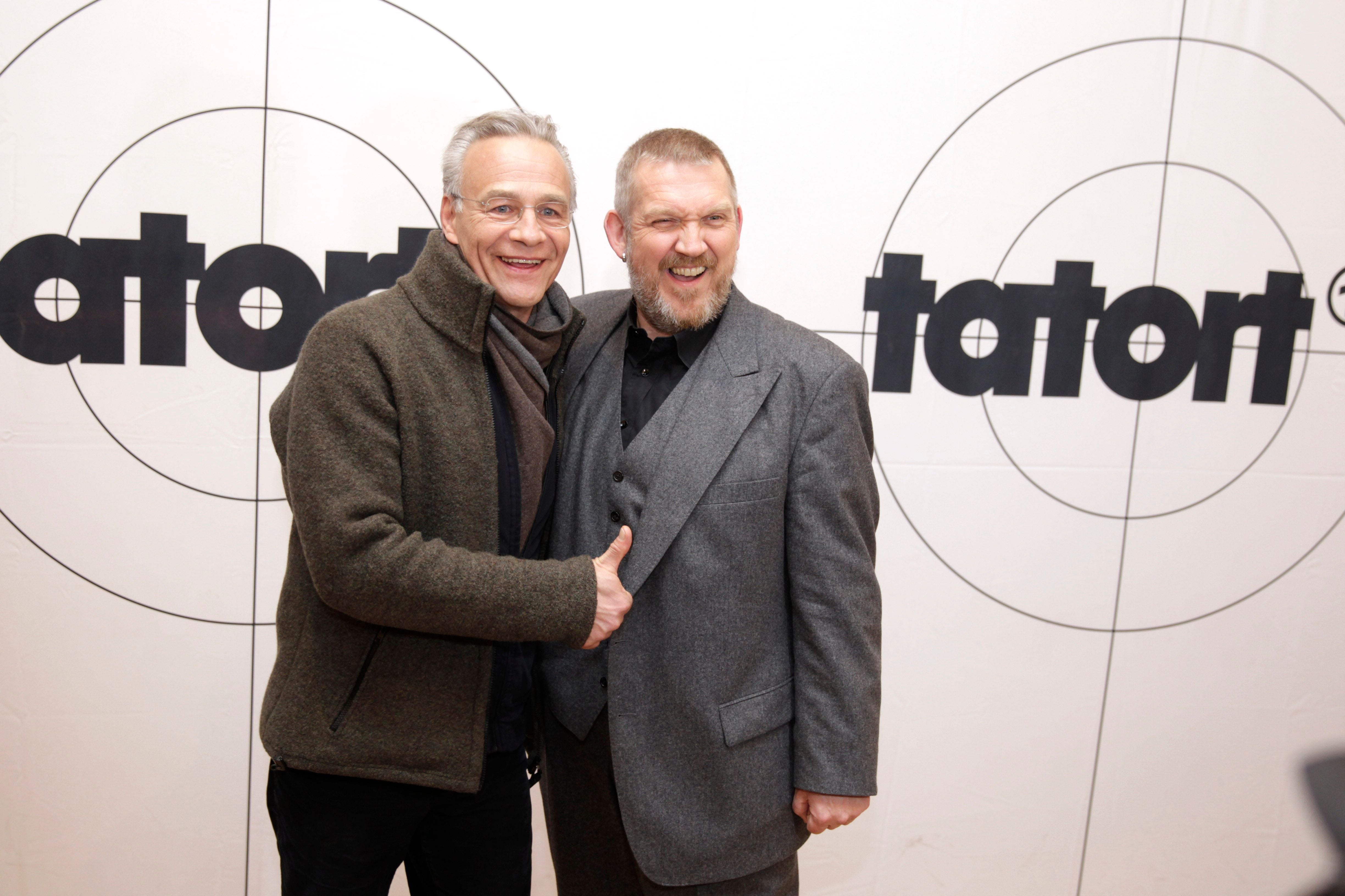 Zwei Männer posieren lachend vor einer Wand mit „Tatort“-Logo und Zielscheibenmotiv.