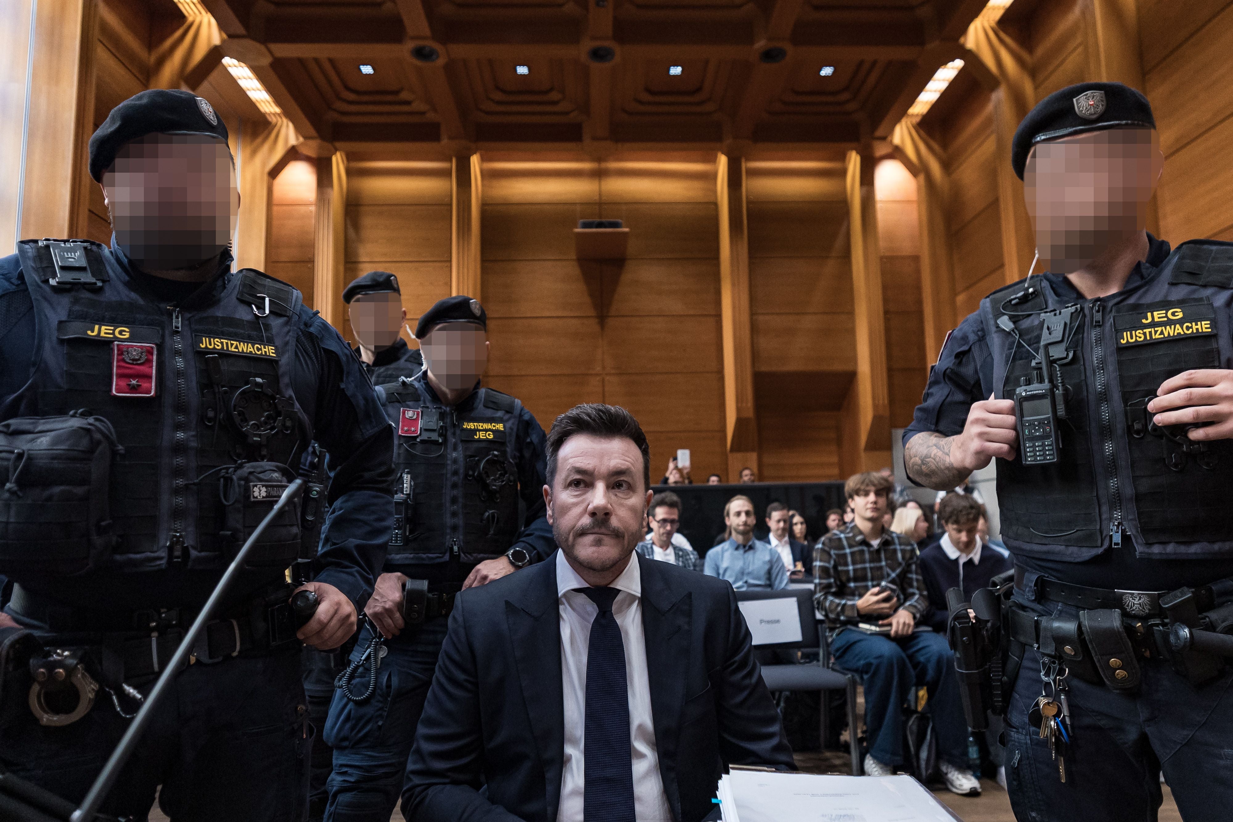 Ein Mann im Anzug sitzt vor vier Justizwachebeamten in einem Gerichtssaal mit Publikum.