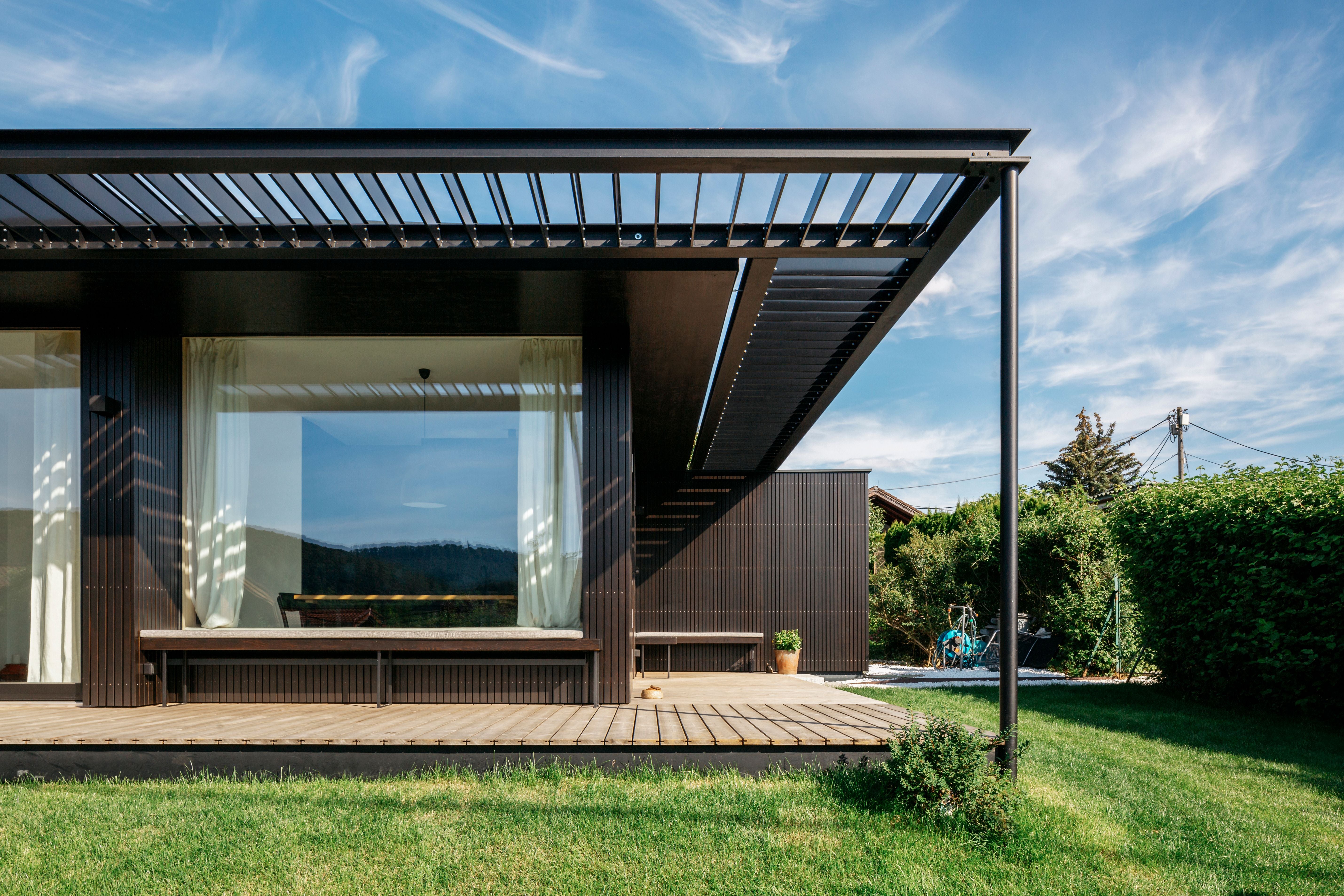 Modernes Tiny House mit Holzterrasse, großen Fenstern und Garten unter blauem Himmel.