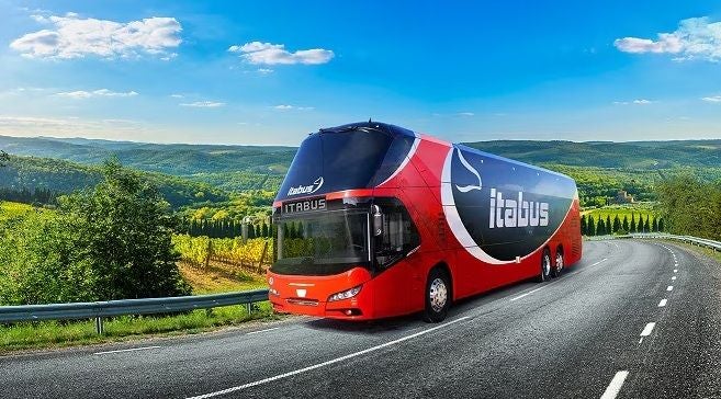 Ein roter Itabus fährt auf einer Landstraße durch eine hügelige, grüne Landschaft.