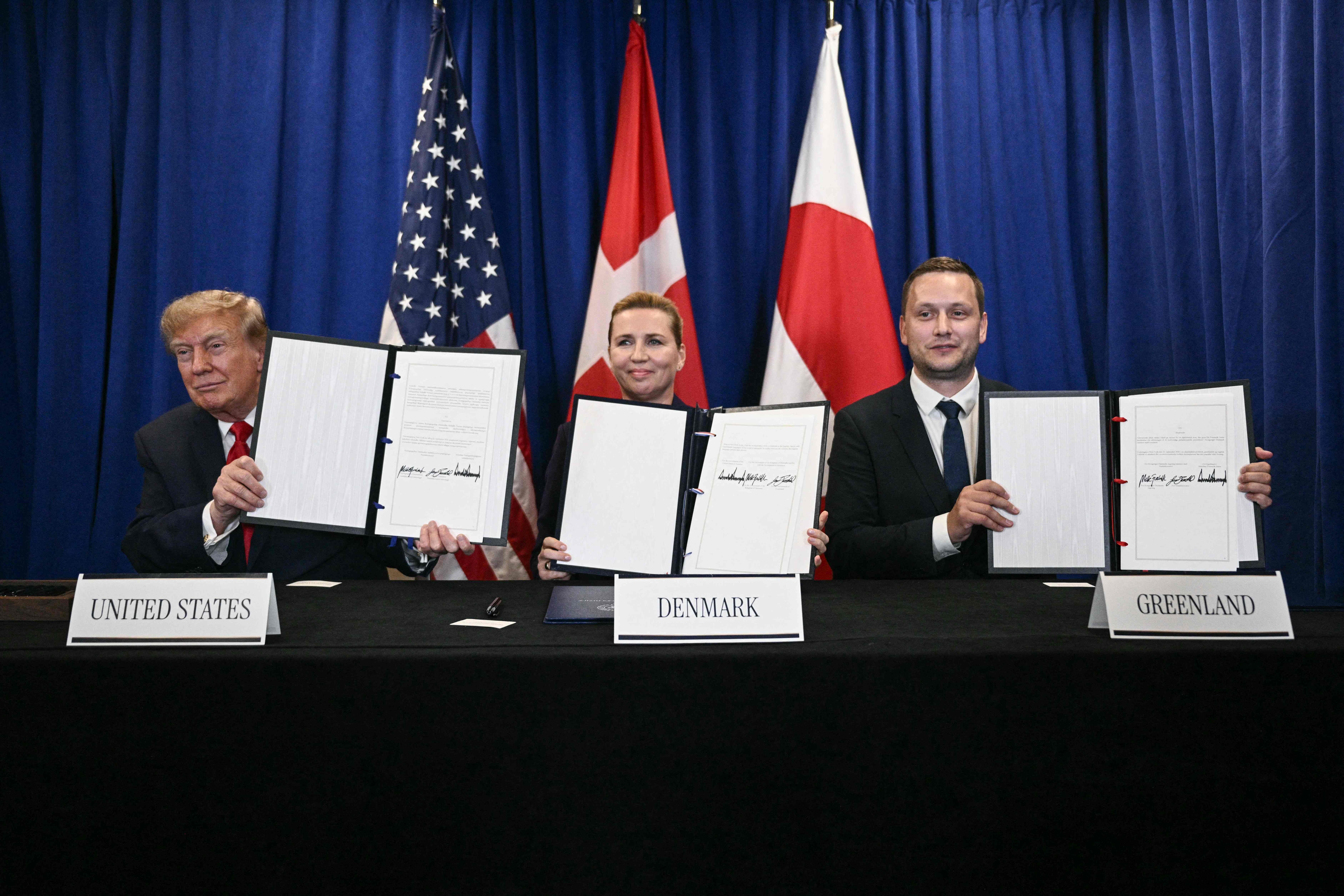 Drei Personen halten unterzeichnete Dokumente vor Flaggen von USA, Dänemark und Grönland hoch.