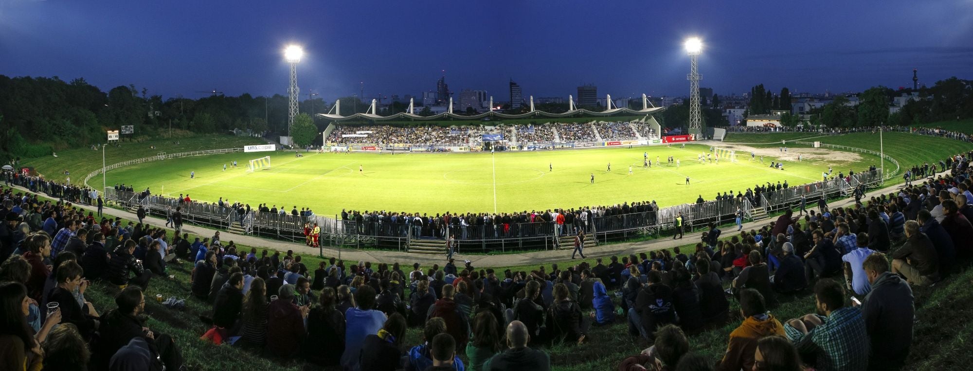 Fußballstadion Hohe Warte abends, viele Zuschauer sitzen auf den Rängen, Flutlicht leuchtet.
