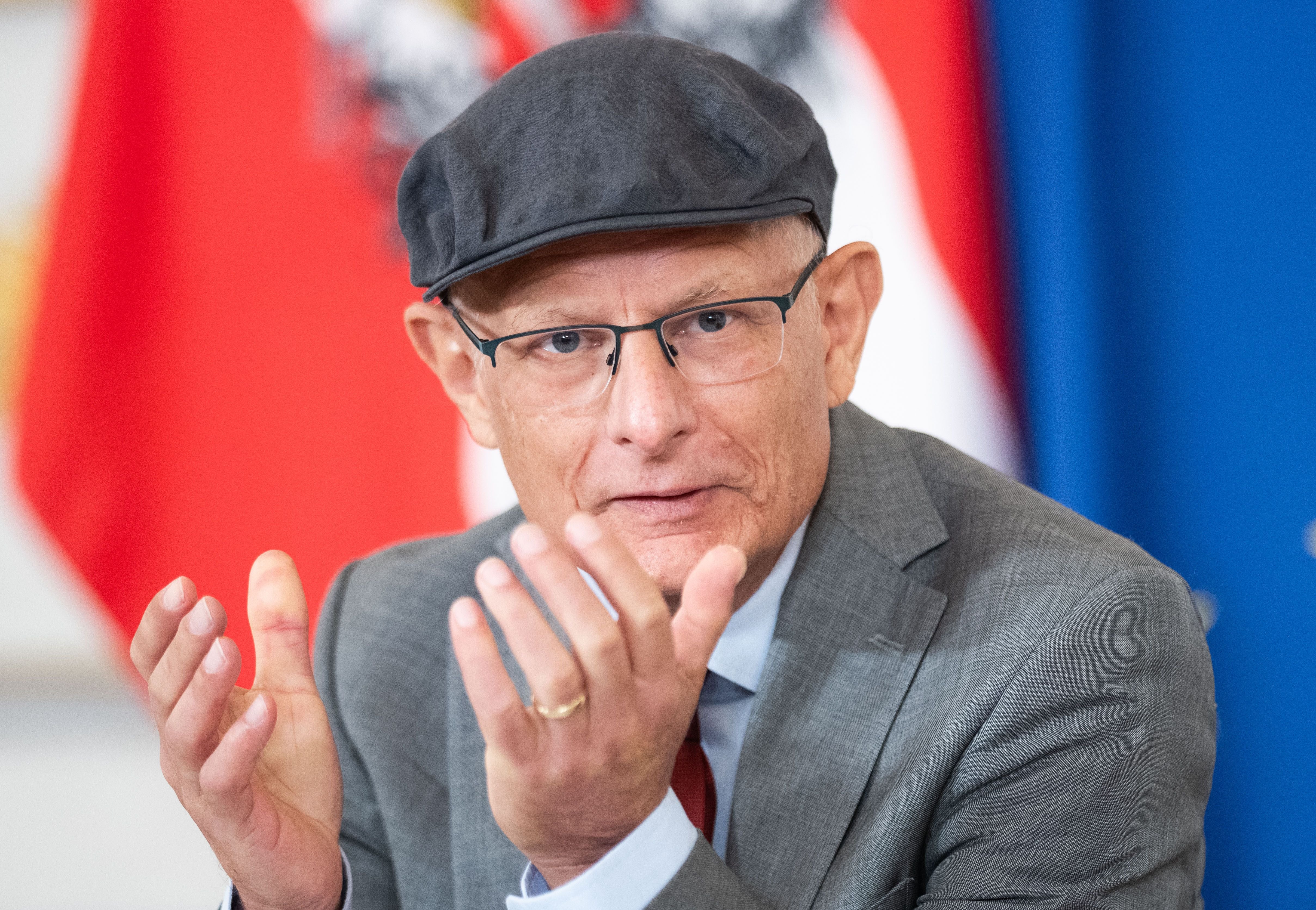 Ein Mann mit Brille und Mütze spricht gestikulierend vor einer rot-weißen Fahne.