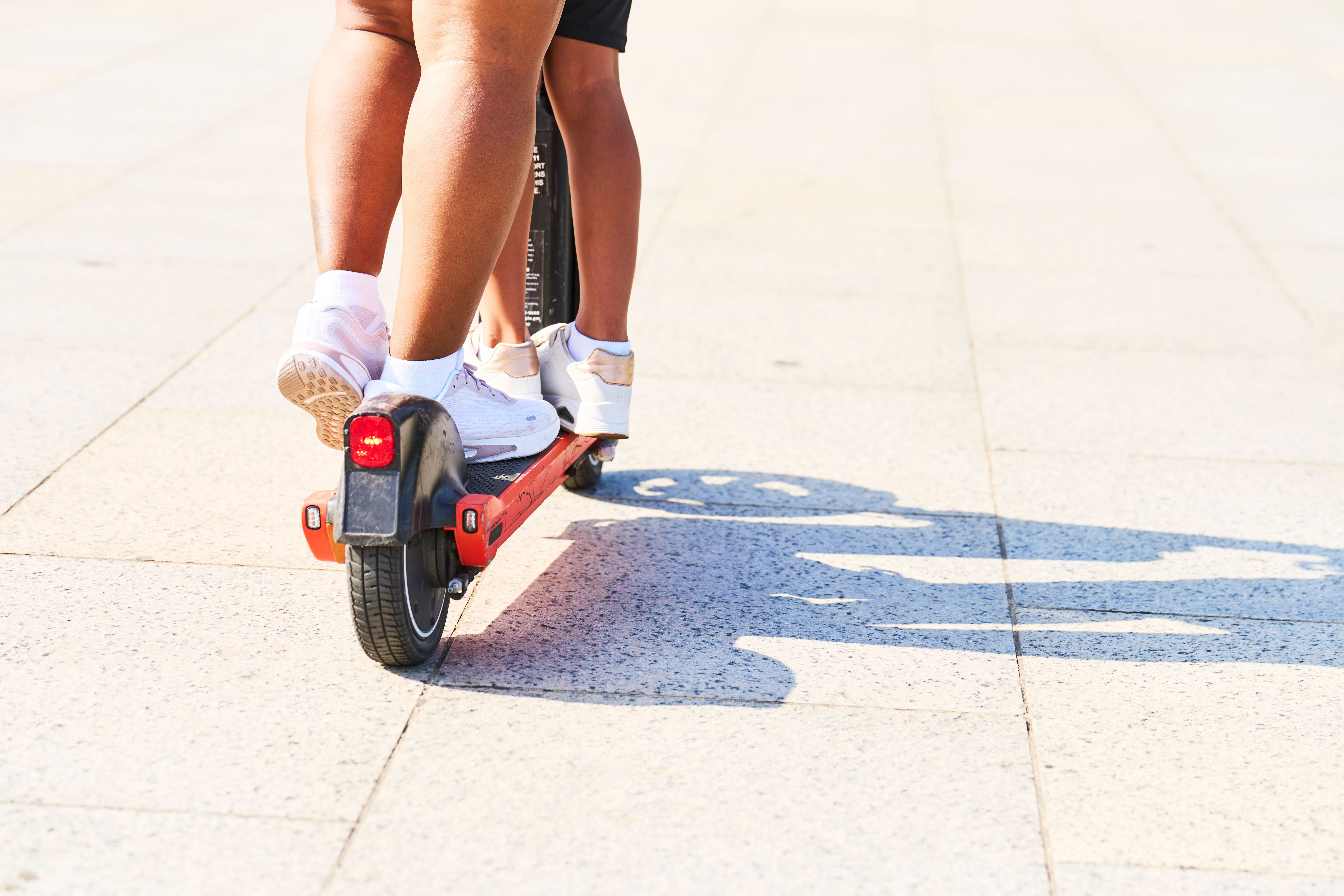 Zwei Personen fahren gemeinsam auf einem E-Scooter auf einem gepflasterten Platz.