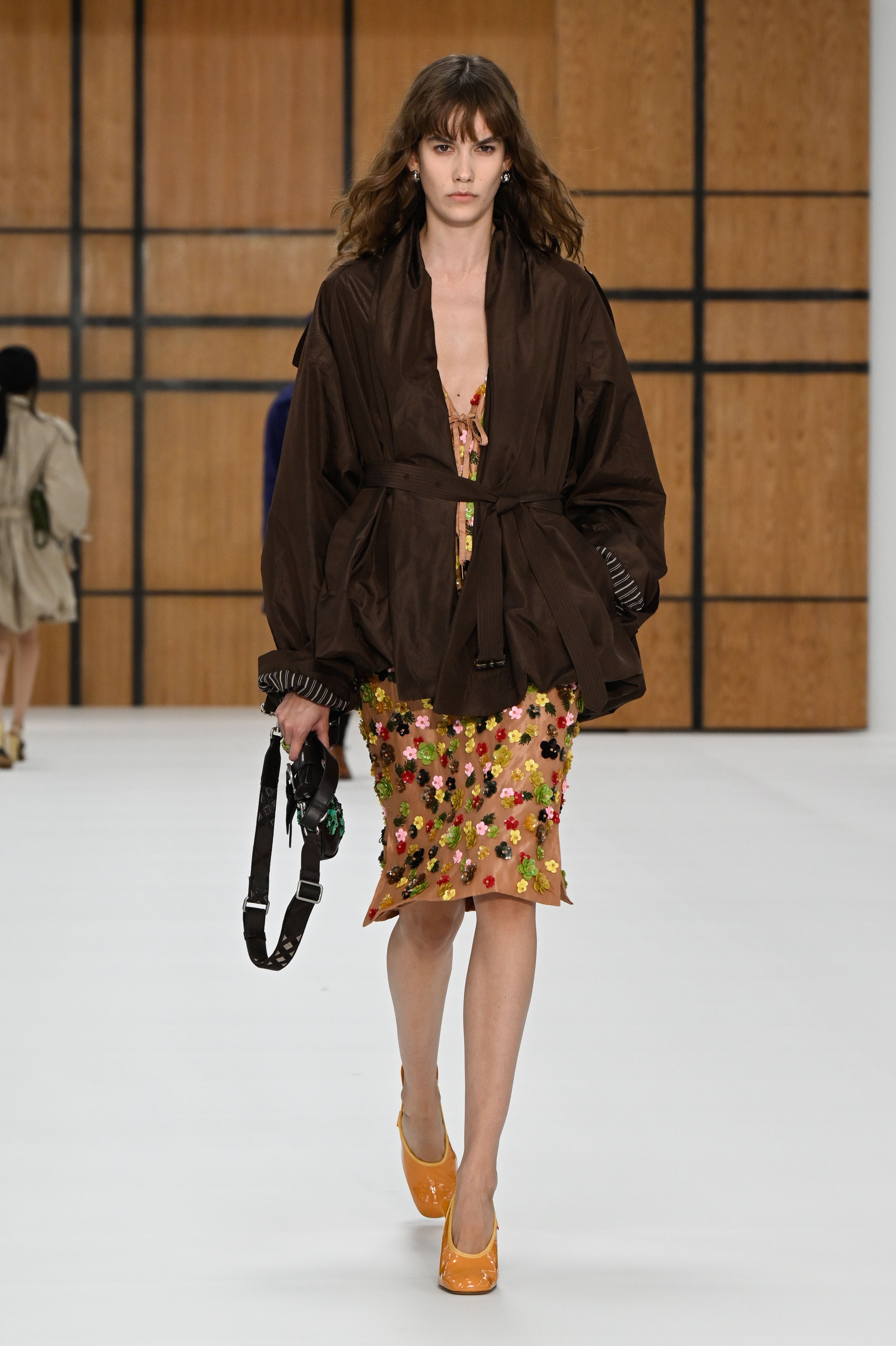 Model mit brauner Jacke, besticktem Rock und gelben Schuhen läuft bei einer Modenschau über den Laufsteg.