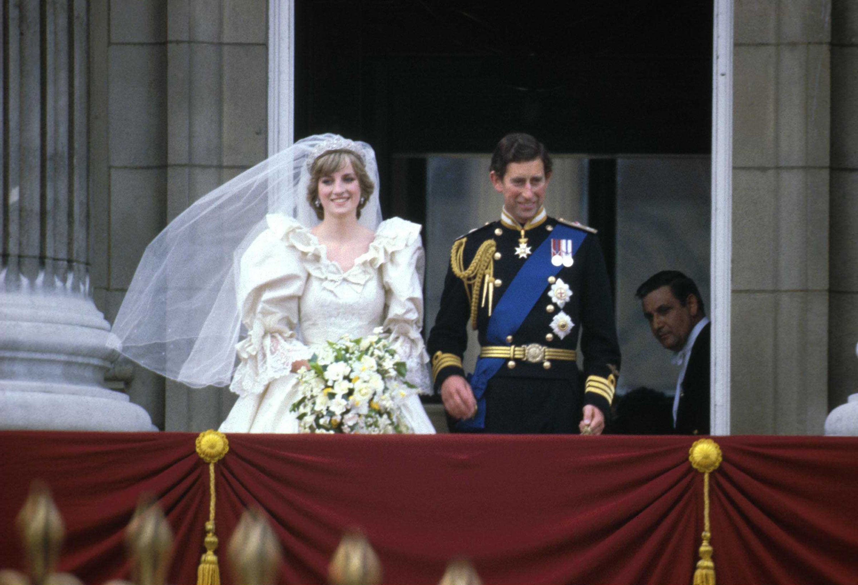 Brautpaar in Hochzeitskleidung steht auf dem Balkon des Buckingham Palace vor rotem Stoff.
