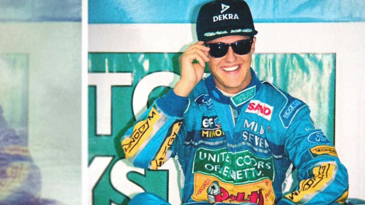 Ein Rennfahrer im blauen Overall lächelt, trägt Sonnenbrille und Kappe mit "DEKRA"-Logo.