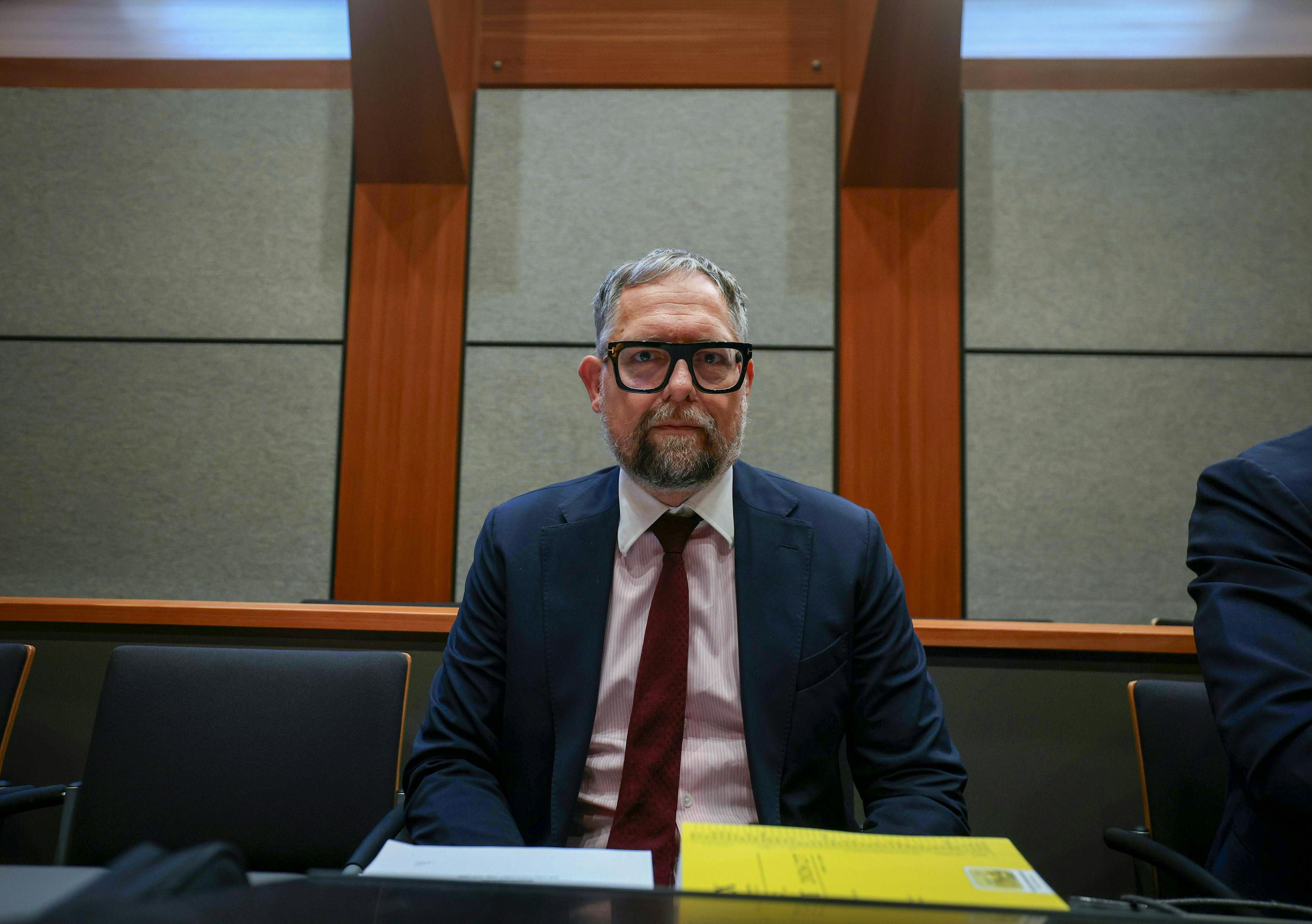 Mann mit Anzug und Brille sitzt in einem Gerichtssaal vor einem Tisch mit Akten.