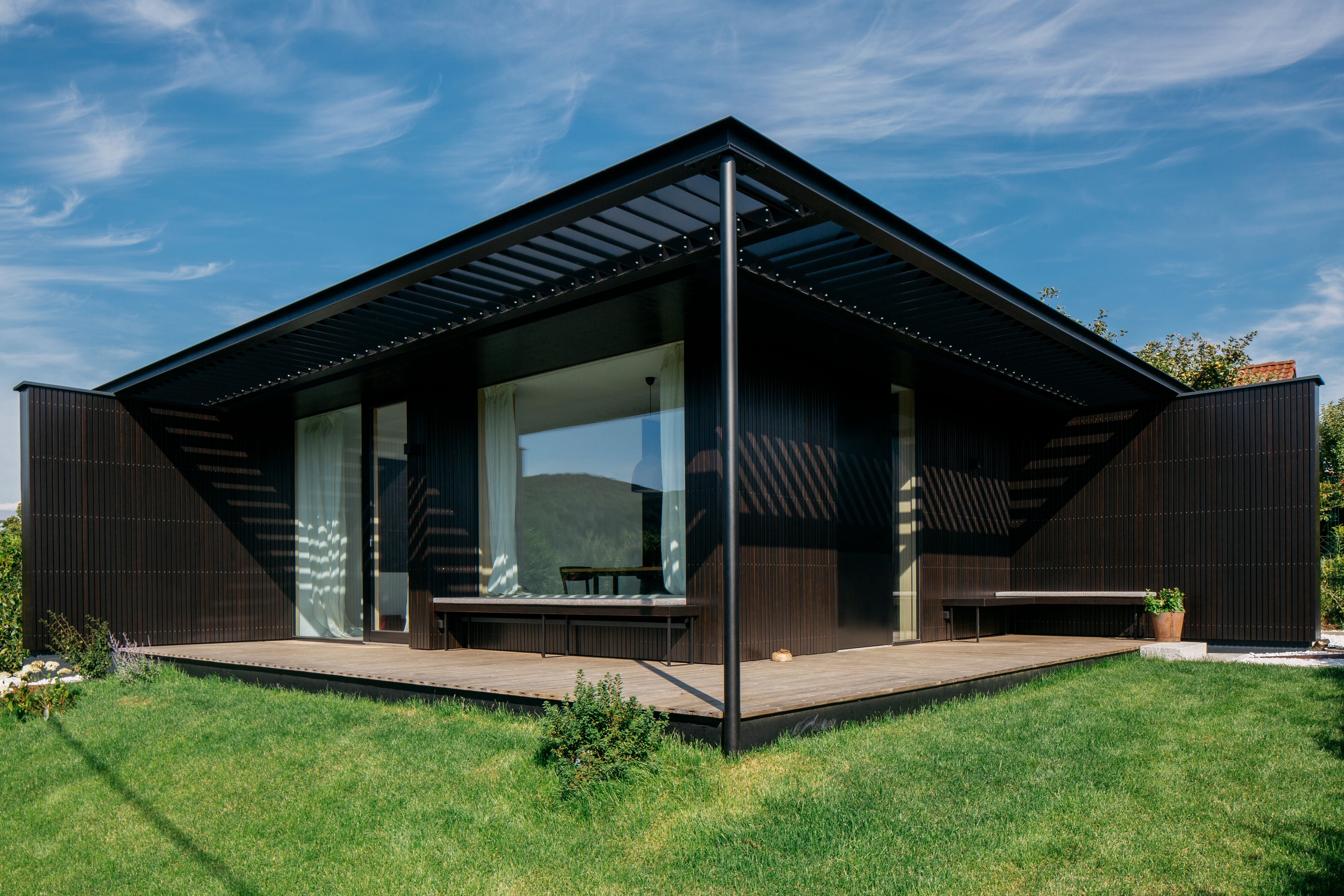 Modernes Tiny House mit dunkler Fassade, großer Terrasse und Glasfront, umgeben von grünem Rasen.
