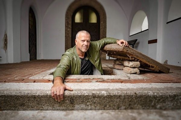 Ein Mann mit grüner Jacke steigt aus einer Bodenluke in einem historischen Gebäude.