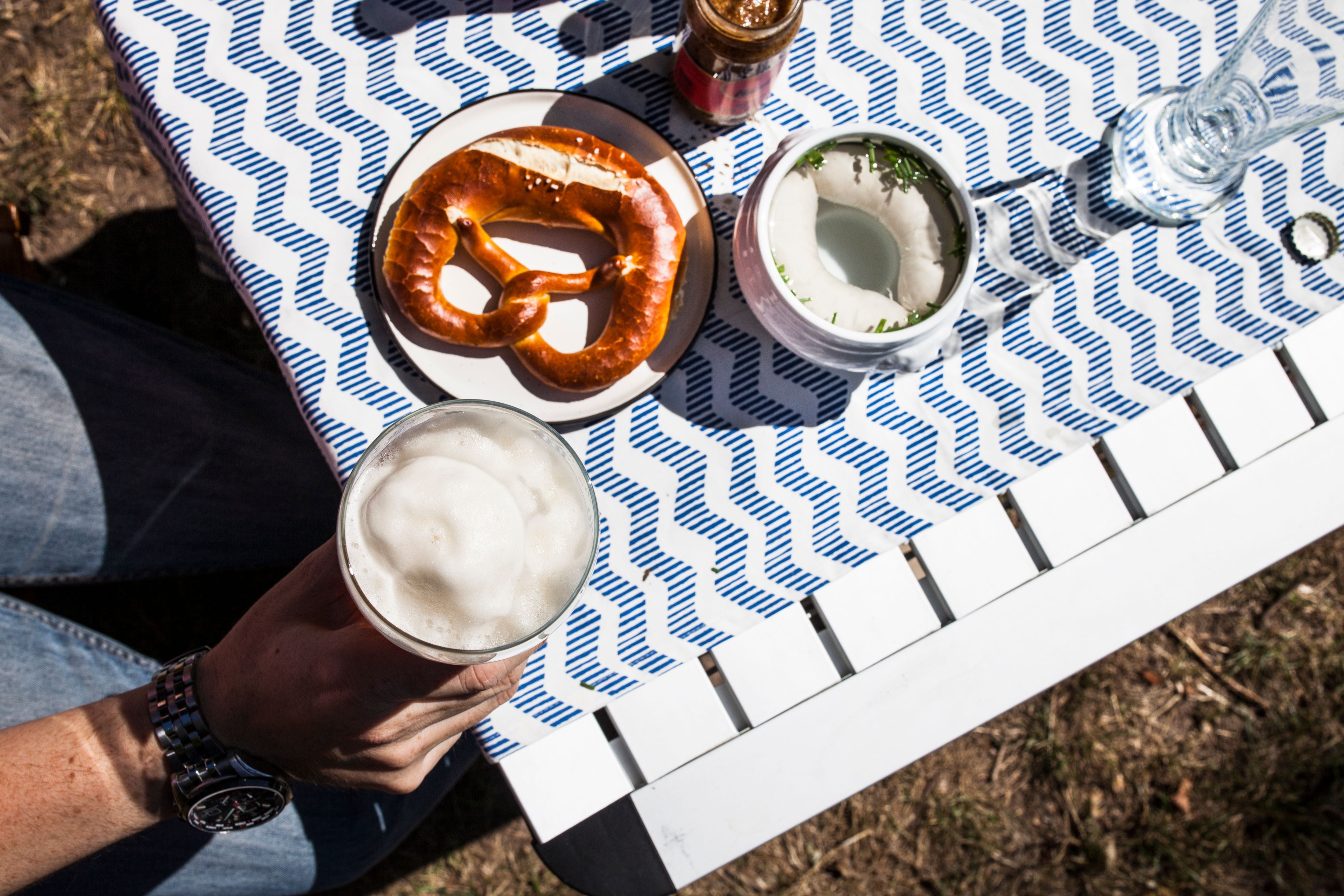 Bierkrug, Brezel und Weißwurst mit Senf auf einem blau-weiß gedeckten Tisch im Freien.