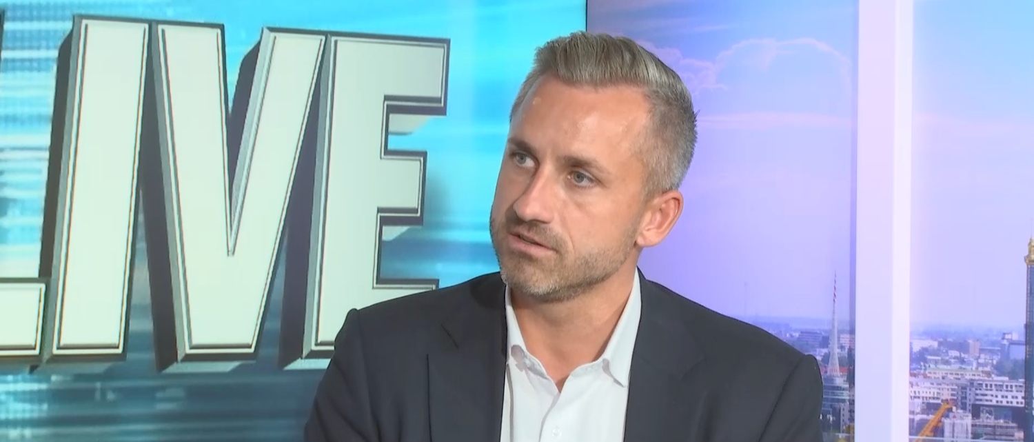 Mann im Anzug spricht im Fernsehstudio mit großem "LIVE"-Schriftzug im Hintergrund.